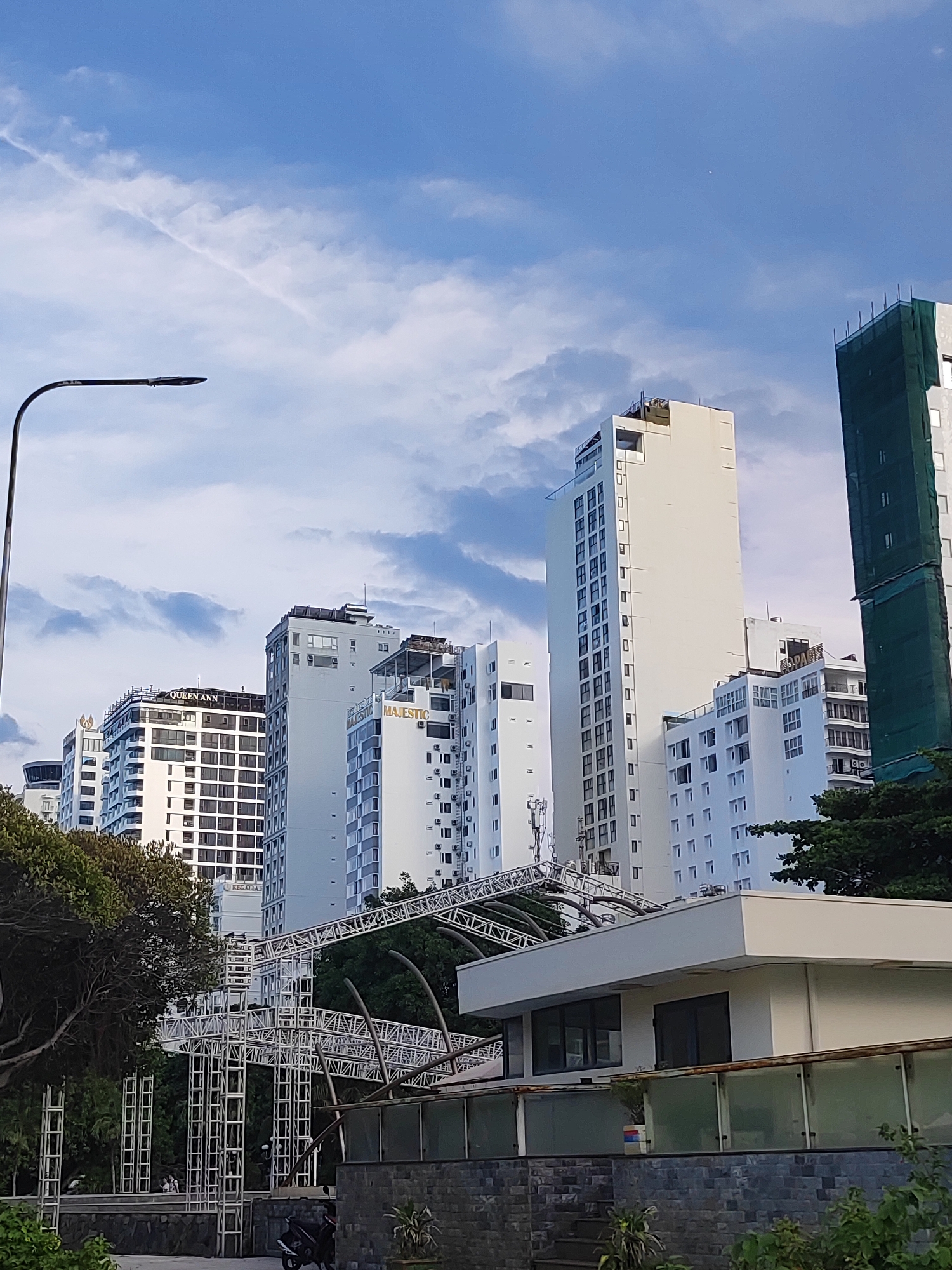 The image depicts a cityscape featuring a row of modern, tall buildings of varying heights, primarily white and light-colored, under a blue sky with scattered clouds. A street lamp with a curved arm is visible on the left side. In the foreground, a white, low-rise building with dark windows is partially visible, along with a stone wall and some greenery. Behind the low-rise building, there is a large, complex metal structure, possibly for an event or construction, with supporting scaffolding. Some of the taller buildings have signs indicating names like "QUEEN ANN" and "MAJESTIC." One tall building on the right is partially covered with green netting, suggesting ongoing construction or renovation. Trees are scattered throughout the scene, adding some natural elements to the urban environment. The overall impression is that of a bustling city area with contemporary architecture and a mix of ongoing activities.