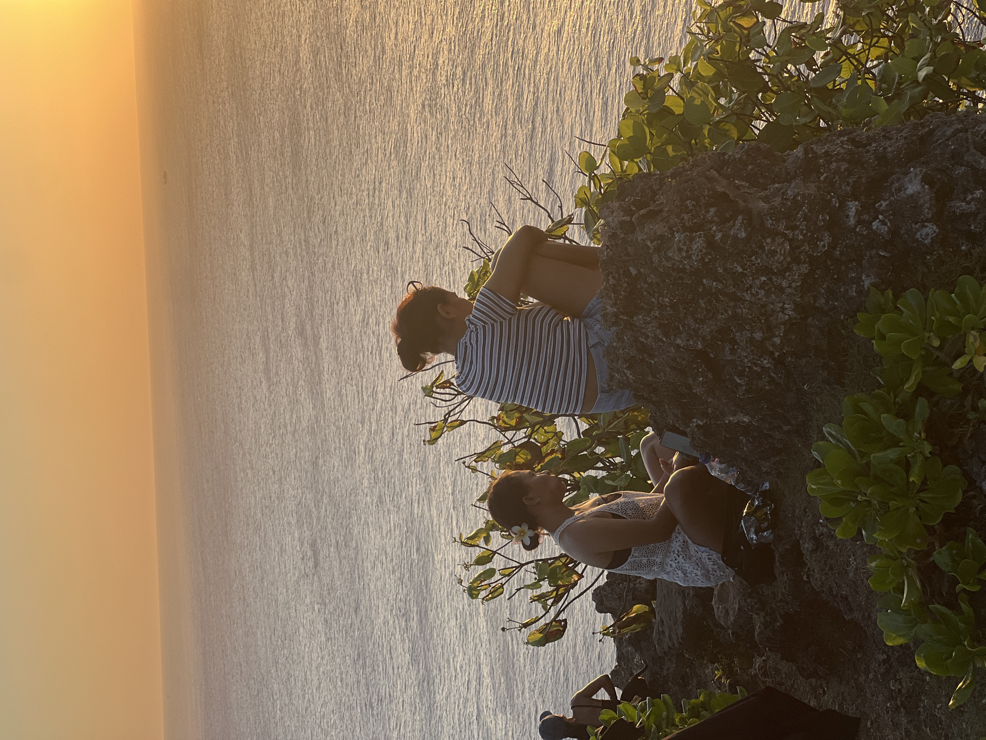 The image depicts a sunset over the ocean in Kuta Selatan, Indonesia, with a group of people gathered on a rocky cliffside. Two women are seated on a large rock in the foreground. The woman on the left is wearing a white lace dress and has a flower in her hair. The woman on the right is wearing a striped t-shirt and denim shorts, with her back to the camera. Several other individuals are visible in the background, some seated on the rocks. Lush green vegetation grows around the rocks. The sky is a warm gradient of orange and yellow, reflecting on the calm ocean surface. A small boat is visible in the distance on the horizon.