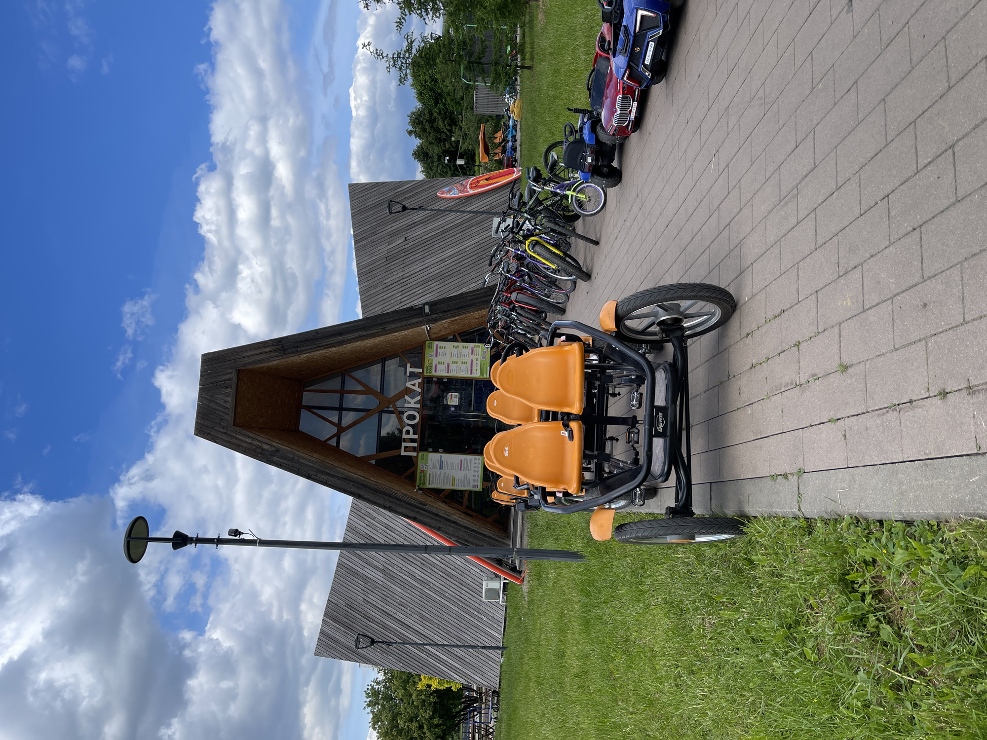 The image shows a recreational area in Moscow, Russia. A rental building, with the Russian word "ПРОКАТ" (rental) prominently displayed, stands with a triangular wooden facade. In front, a four-person pedal bike with orange seats is parked on a paved path bordering a grassy area. Various bicycles and small electric cars are parked nearby. The sky is partly cloudy, and a lamppost stands to the left of the rental building.