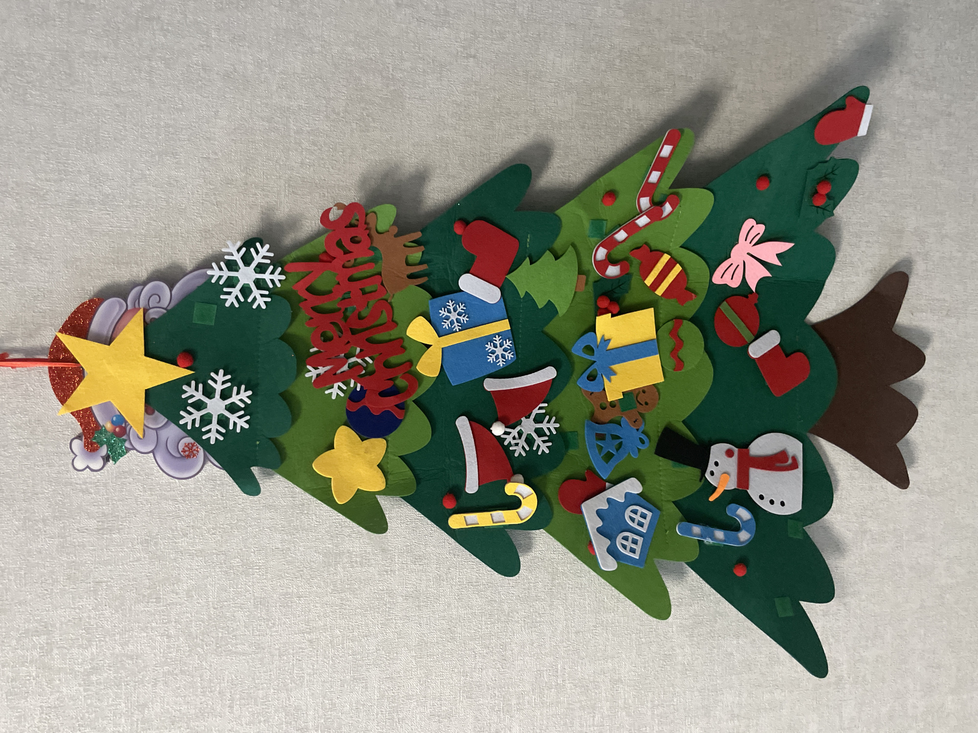 A felt Christmas tree decoration is on a light gray surface. The tree is green with a brown trunk and decorated with felt ornaments including stars, snowflakes, gifts, candy canes, a snowman, a house, and other festive items. The ornaments are colorful and are attached to the tree. The image does not show any information that it is located in the city of Izhevsk, Russia.