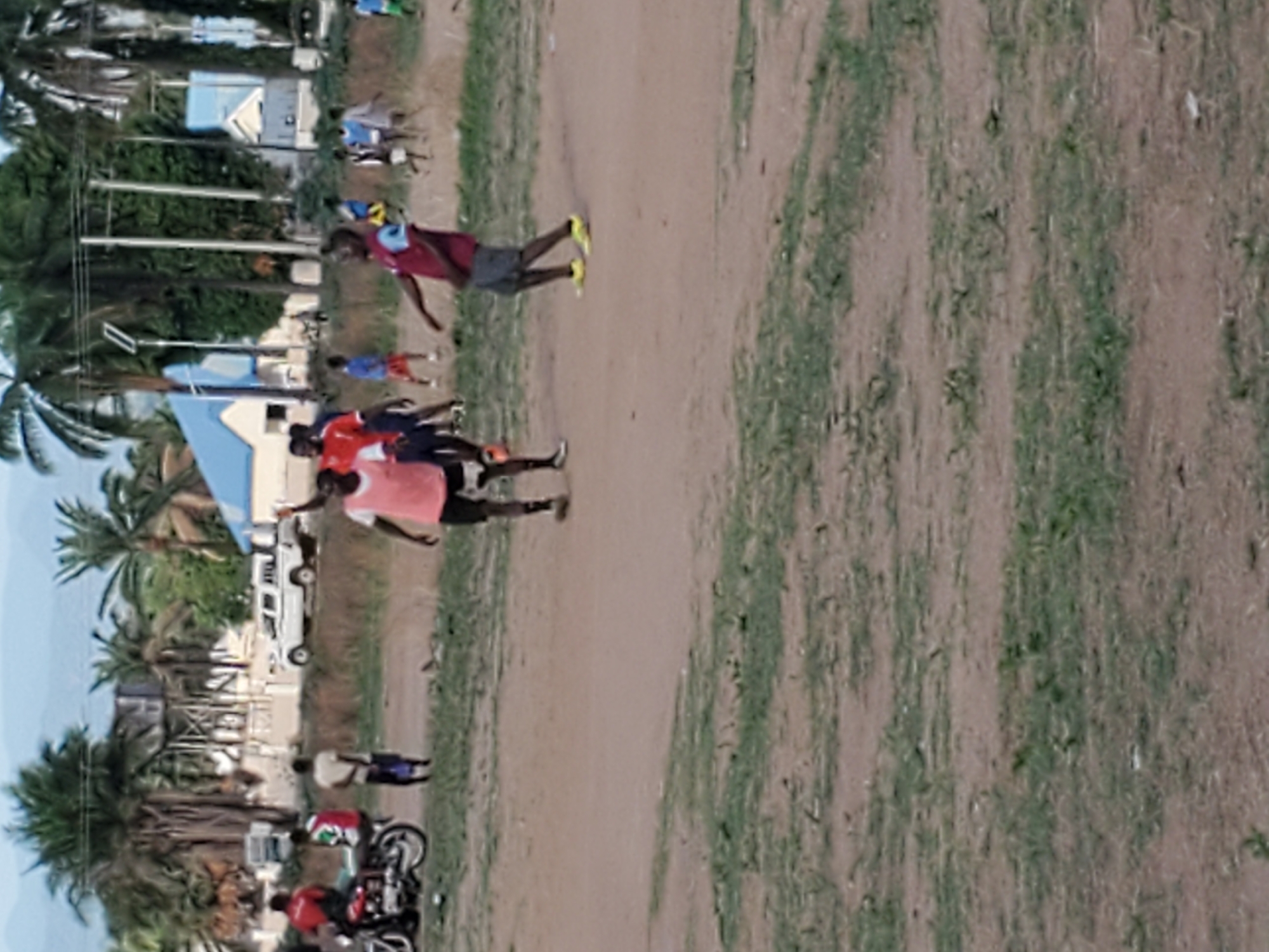 This outdoor image, taken in what appears to be Mararaba, Nigeria, depicts an open, unpaved area with scattered individuals and background structures under bright daylight. The ground is a mix of light brown dirt and sparse patches of green grass.
In the midground, multiple people are present. A person wearing a maroon-red shirt, dark shorts, and distinctive neon yellow athletic shoes is captured in a dynamic pose, as if walking or in motion, on the dirt path. To their right, a small group includes an individual in a red shirt and another in a pinkish-orange shirt, appearing to be conversing or observing. Other figures in various colored shirts (including blue) are spread across the scene.
The background is rich with tropical foliage, primarily featuring numerous tall palm trees and other dense green trees. Behind the vegetation, several buildings with light-colored walls and bright blue roofs are visible, suggesting local architecture. Utility poles stand amongst the trees, indicating infrastructure. A white vehicle, possibly a pickup truck or SUV, is seen in the mid-background near the buildings. On the far right, a person in a red shirt stands beside a motorcycle.
The lighting suggests a sunny, clear day. The overall impression is one of casual daily activity, possibly a gathering or transit through an open community space. No legible text is visible in the image.