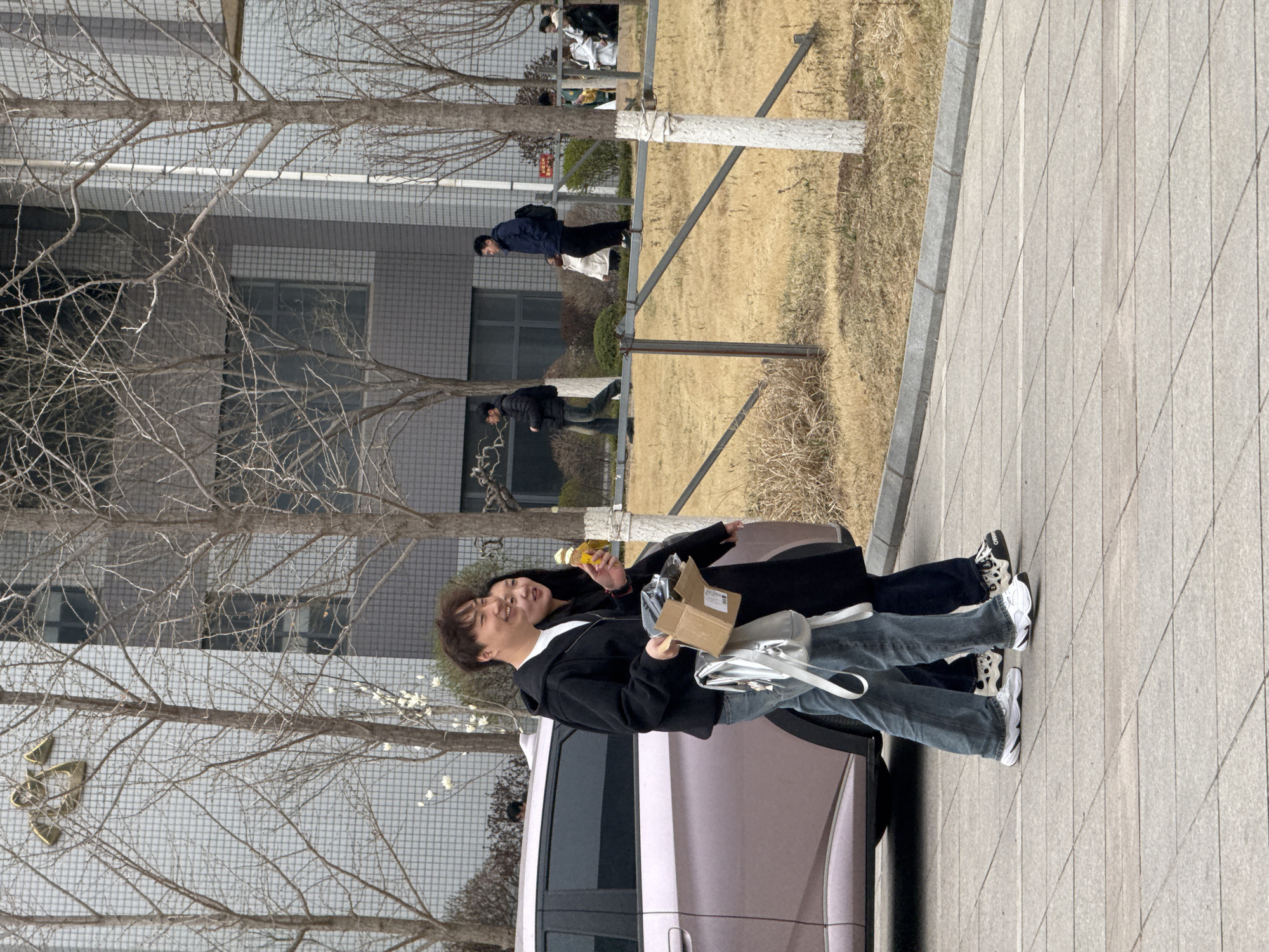 Two people stand on a paved sidewalk next to a car. One person, on the left, holds a small cardboard box and a silver handbag. The other person holds a bottle. Behind them is a car, and further back, buildings and bare trees. In the distance are a grassy area and other people walking. The location is identified as Zhongtun, China.
