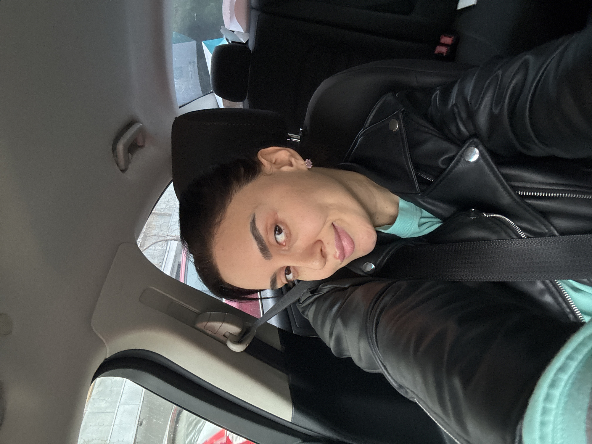 A medium shot, rotated 90 degrees clockwise, captures a woman taking a selfie from inside a car, likely in Tbilisi, Georgia. The woman, with dark hair pulled back, gazes directly at the camera with a calm expression and a slight smile. She is wearing a black leather jacket with silver snap buttons and zippers, over a light blue or mint green long-sleeved top. A small, light-colored (possibly pink or purple) stud earring is visible in her left ear.
She is seated in the passenger seat, with a black seatbelt fastened across her chest. The car's interior is visible, featuring dark seats and a light grey headliner and pillar on the left side of the frame, where a small handle or hook is present. Through the car window, hints of an urban environment with buildings and possibly concrete structures can be seen, suggesting a city setting during daytime. The lighting appears natural and bright, indicating it's not night or heavily overcast. No visible text is present in the image.