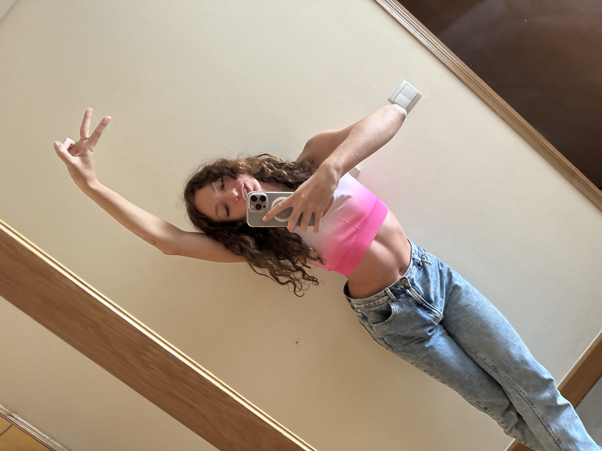 A young woman is taking a selfie in front of a mirror. She is wearing a pink and white tie-dye top and blue jeans. She is making a peace sign with her left hand. The background is a wall with a light switch and a wooden trim. There is no indication of Đà Nẵng, Vietnam.