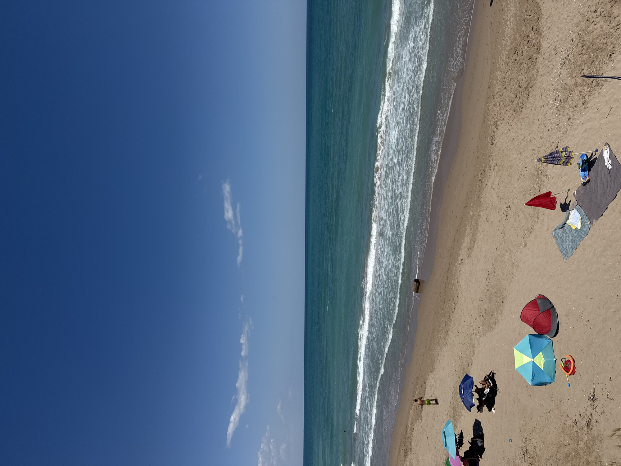 The image depicts a sandy beach on a sunny day with a clear blue sky. The turquoise sea stretches to the horizon, with gentle waves breaking on the shore. In the foreground, several beach umbrellas and tents are set up, providing shade. There are also towels and beach toys scattered on the sand. A person is standing near the water's edge, and another person is lying under an umbrella in the middle ground. The overall scene suggests a relaxed day at the beach, likely in the vicinity of Shabla, Bulgaria, known for its coastal resorts.