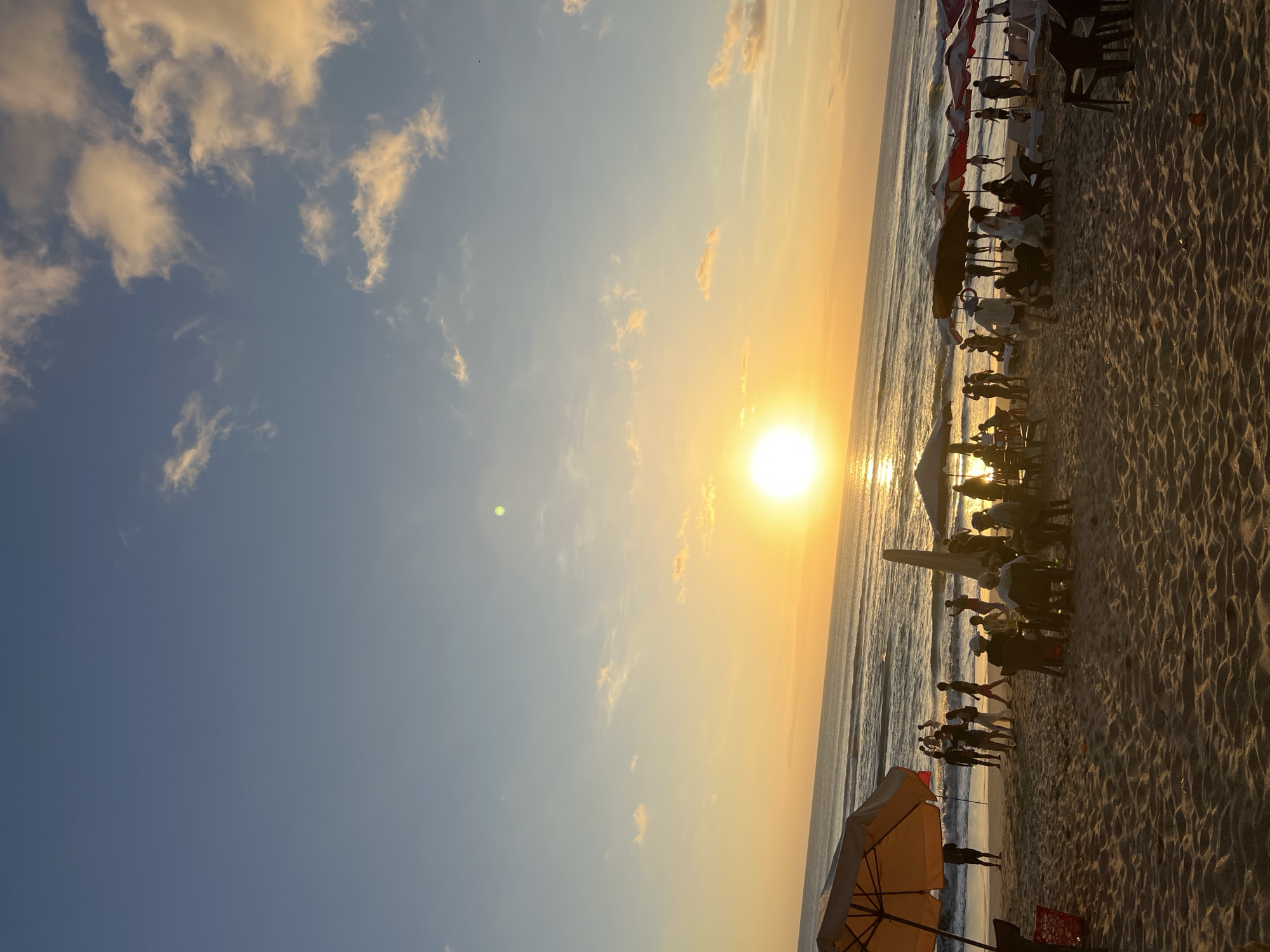 The image depicts a beach scene in Kuta, Indonesia, during sunset. The sun is a bright orb low on the horizon, casting a warm golden light across the sky and reflecting on the ocean waves. The sky is a gradient of blue with scattered clouds tinged with orange and yellow from the setting sun.
On the sandy beach, there are several beach umbrellas, some open and providing shade, others closed. People are scattered across the beach, some walking, some sitting on chairs, and others gathered in small groups. A few individuals are visible in the shallow water at the edge of the ocean. The overall atmosphere suggests a relaxed evening gathering on the beach.
The foreground is dominated by the textured sand of the beach, with some chairs and a discarded red basket visible. The middle ground features a cluster of people near umbrellas and beach chairs, with more figures dotted further down the beach towards the water. The background consists of the ocean and the horizon line where it meets the sky. The presence of numerous people enjoying the sunset indicates a popular public space.