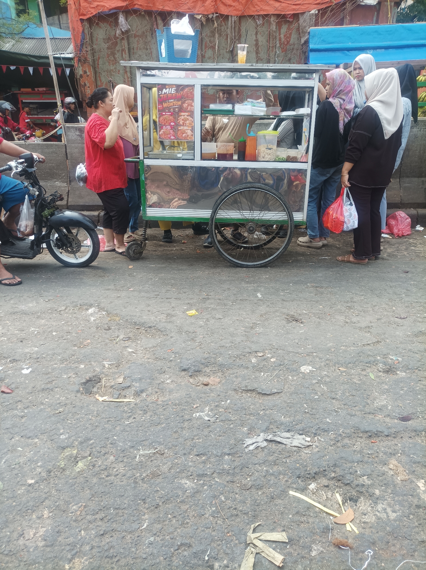 The image captures a street scene in South Jakarta, Indonesia, focusing on a food cart and several individuals. A stainless steel food cart, likely selling noodles and wontons as indicated by signage, is the central element. The cart has a glass display case and two large wheels at the back, with smaller stabilizing wheels at the front. Several people are gathered around the cart, some appearing to be customers and others possibly vendors. One woman in a red shirt is looking towards the cart, while others are partially obscured or engaged in transactions. A motorcycle is parked to the left of the frame, with its front wheel visible. The ground is paved and appears worn, with some debris scattered on its surface. The background shows a covered structure with orange tarpaulin and a glimpse of buildings and vegetation, suggesting an urban environment. The lighting indicates it is daytime.