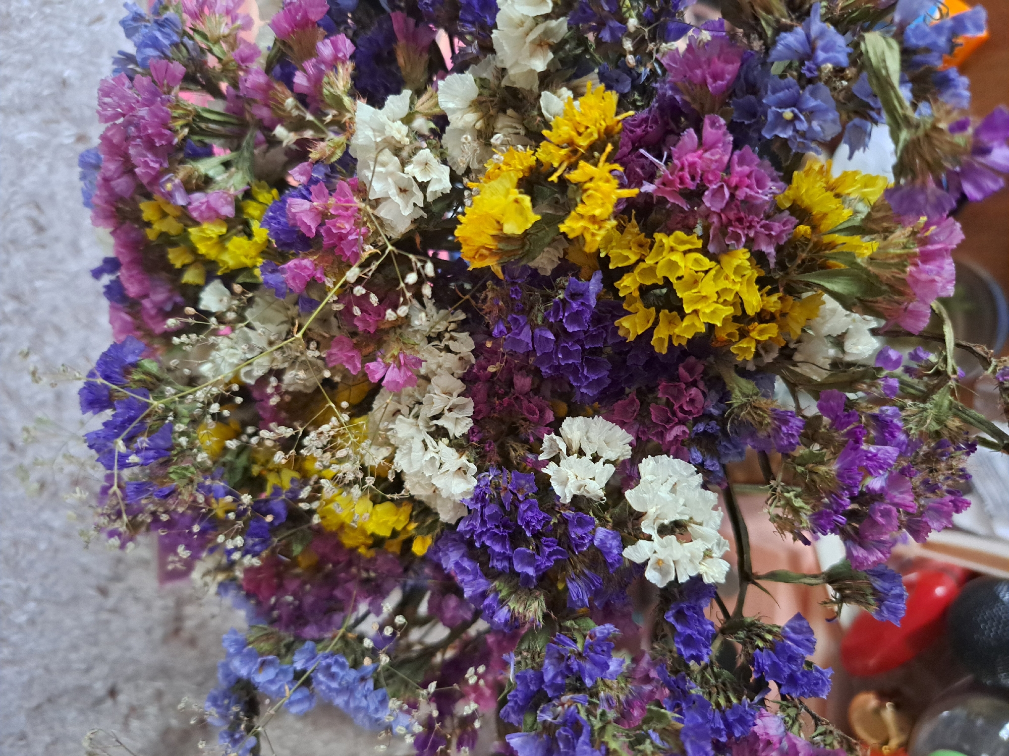 A close-up, eye-level shot captures a vibrant bouquet of dried flowers. The bouquet is densely packed and fills the frame, showcasing a variety of colors including purple, blue, pink, yellow, and white. The flowers appear to be statice, known for their papery texture and long-lasting nature. The background is out of focus and appears to be a textured wall, possibly a building exterior, suggesting the image was taken outdoors or near an entrance. In the lower portion of the frame, out-of-focus objects include what appear to be the tops of bottles or containers and a red object, possibly a plastic toy or decorative item. The lighting is bright, casting a soft glow on the colorful blooms. No people are visible in the scene. This image is unlikely to be specifically located in the city of Vypasne, Ukraine, as there are no identifying landmarks or contextual clues to suggest this location.