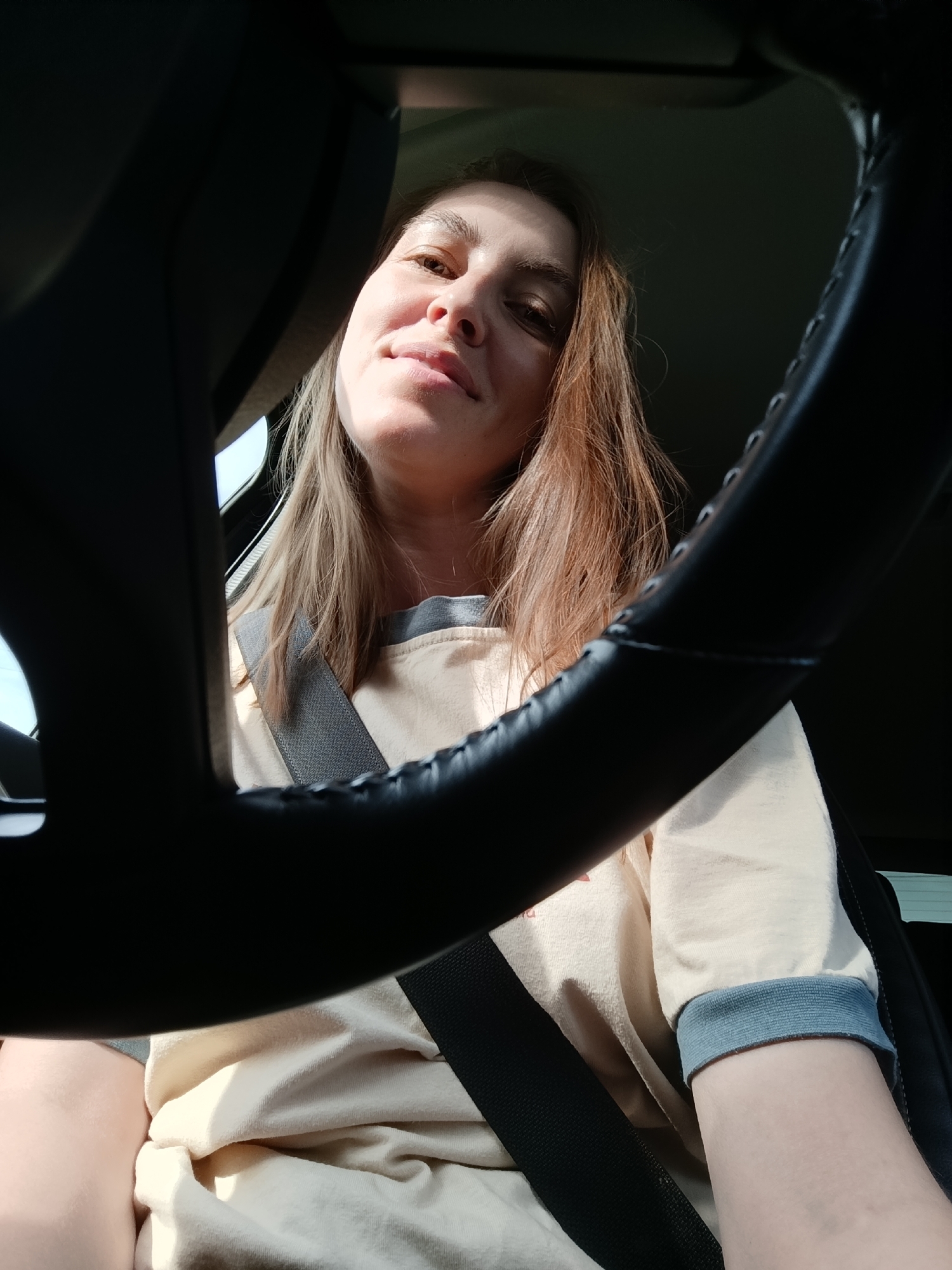 A woman is in the driver's seat of a car. She has blonde hair and is wearing a beige shirt with blue cuffs. She is wearing a seatbelt. The steering wheel is in the foreground. The location is unknown, including the city of Chaikovsky, Russia.