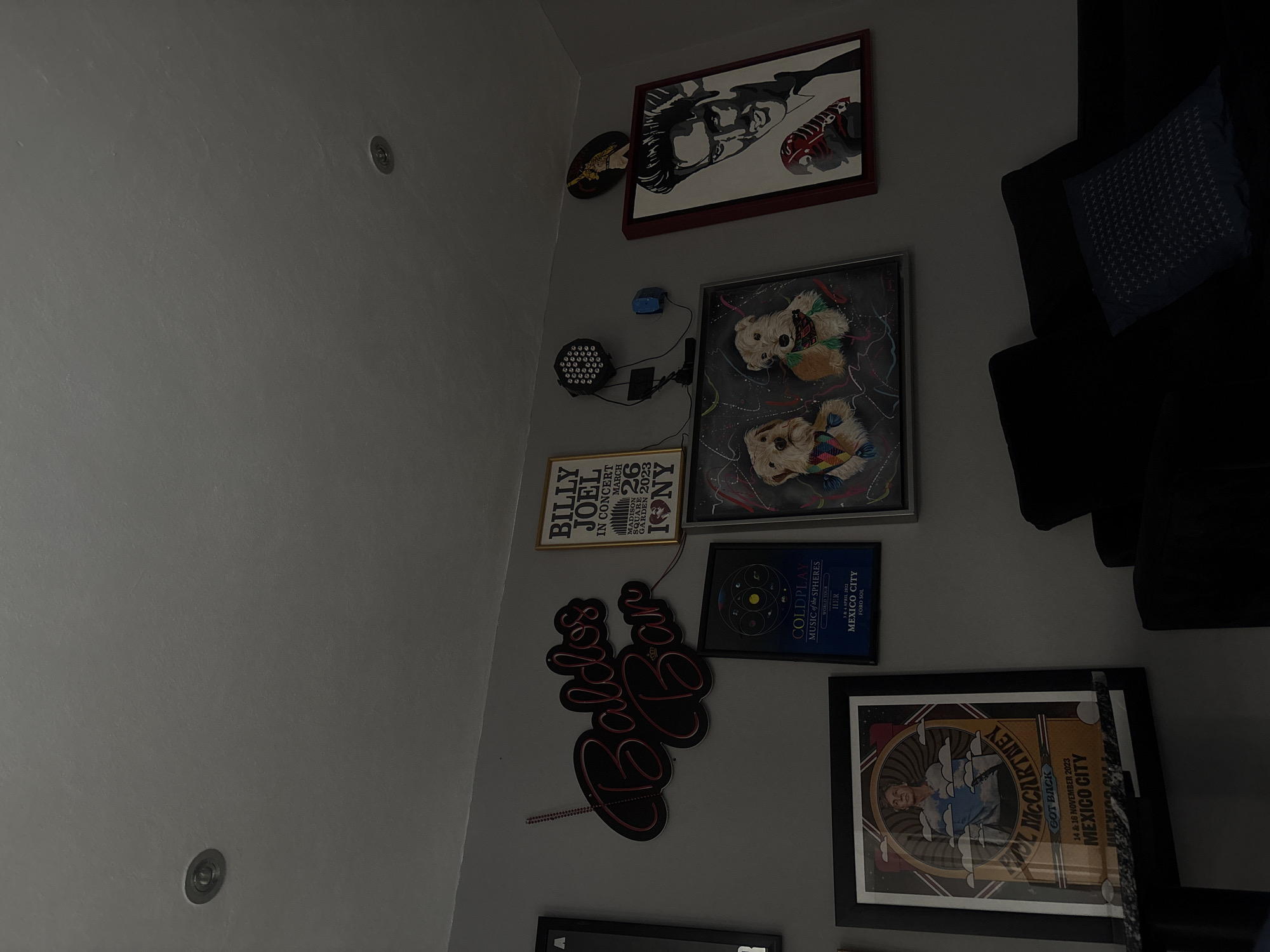 The image shows a wall with several framed posters and decorations. The posters include a Billy Joel concert poster from Madison Square Garden on March 26, 2023, a Paul McCartney "Got Back" tour poster from Mexico City on November 14 & 16, 2023, and a Coldplay "Music of the Spheres" world tour poster from Mexico City on April 3 & 4, 2022. There is also a framed picture of two dogs and a black and white artwork in a red frame. A neon sign reading "Coldest Bar" is visible. There is a light fixture with multiple small lights attached to the wall. A piece of a dark-colored couch with a patterned pillow is visible on the right side of the frame. The wall is gray, and there are two ceiling lights visible on the left side. The location in San Bernardino Tlaxcalancingo, Mexico, is not mentioned.