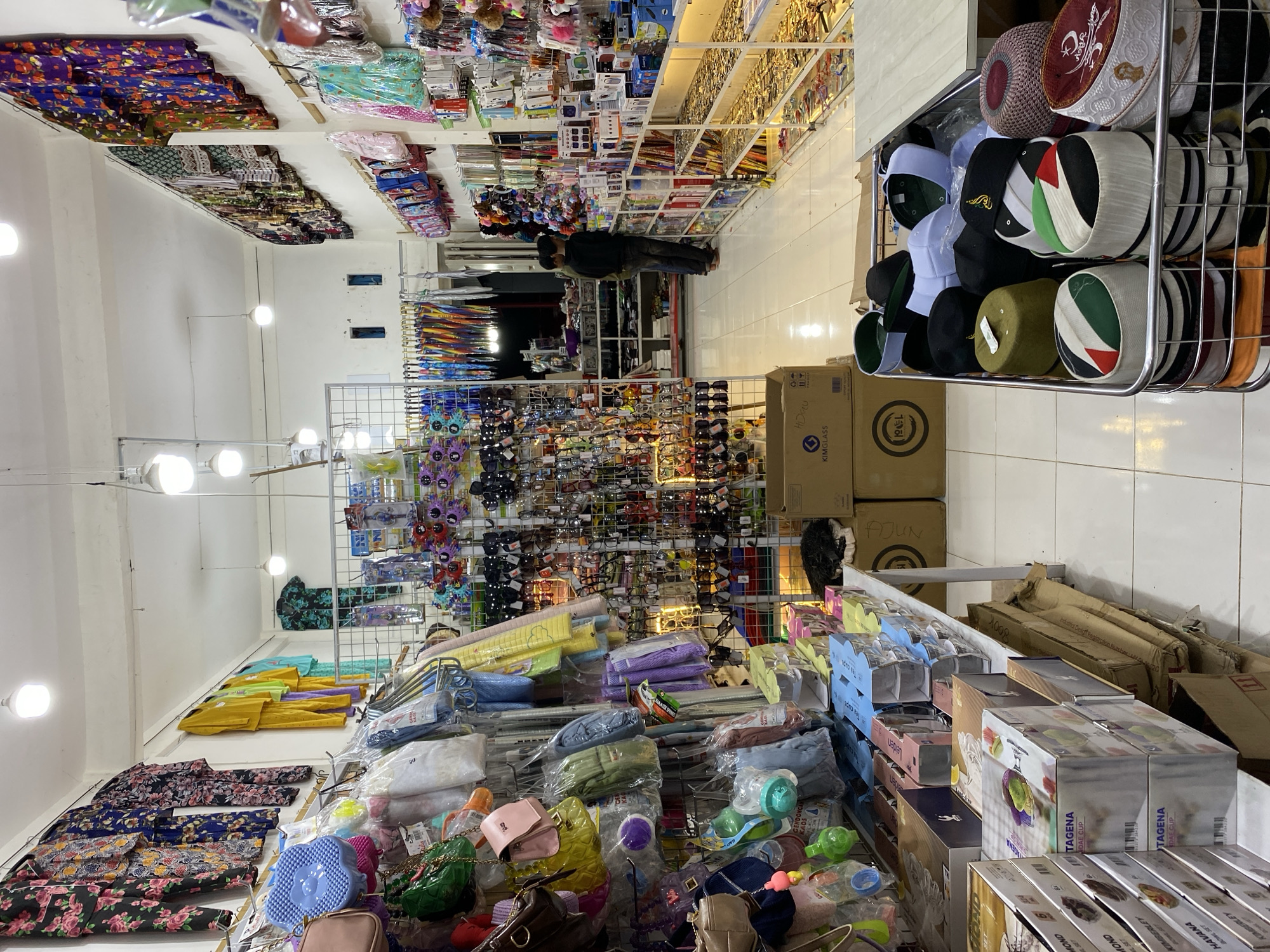 The image shows the interior of a small store in Lamteumen Barat, Indonesia, filled with a variety of merchandise. The store is brightly lit by overhead fluorescent lights. Racks and shelves are densely packed with items, including clothing, accessories, and packaged goods.
On the left side of the image, hanging clothing and accessories are displayed, such as floral patterned pajamas, colorful tops, handbags, and smaller items like keychains and decorative objects. Further back, there are racks of what appear to be umbrellas and more hanging items.
In the center of the store, a large wire rack contains an assortment of hats, including skullcaps and kufi hats in various colors and designs. Some of these hats are decorated with national flags, such as the Palestinian flag. Adjacent to the hat display are boxes of packaged goods, possibly kitchenware or home goods, stacked on shelves and a table.
On the right side of the store, shelves display more merchandise, including what look like electronic accessories or phone cases, as well as more general retail items. Further back, there is a glimpse of another section of the store with more shelving.
Two individuals, appearing to be men, are standing in the middle-right section of the store, looking at the merchandise on the shelves. One is wearing a black long-sleeved shirt and dark pants, and the other is wearing a dark olive green shirt and dark pants. They appear to be customers or employees.
The flooring is tiled, and the walls are painted a light color. The overall impression is of a busy, well-stocked retail space.