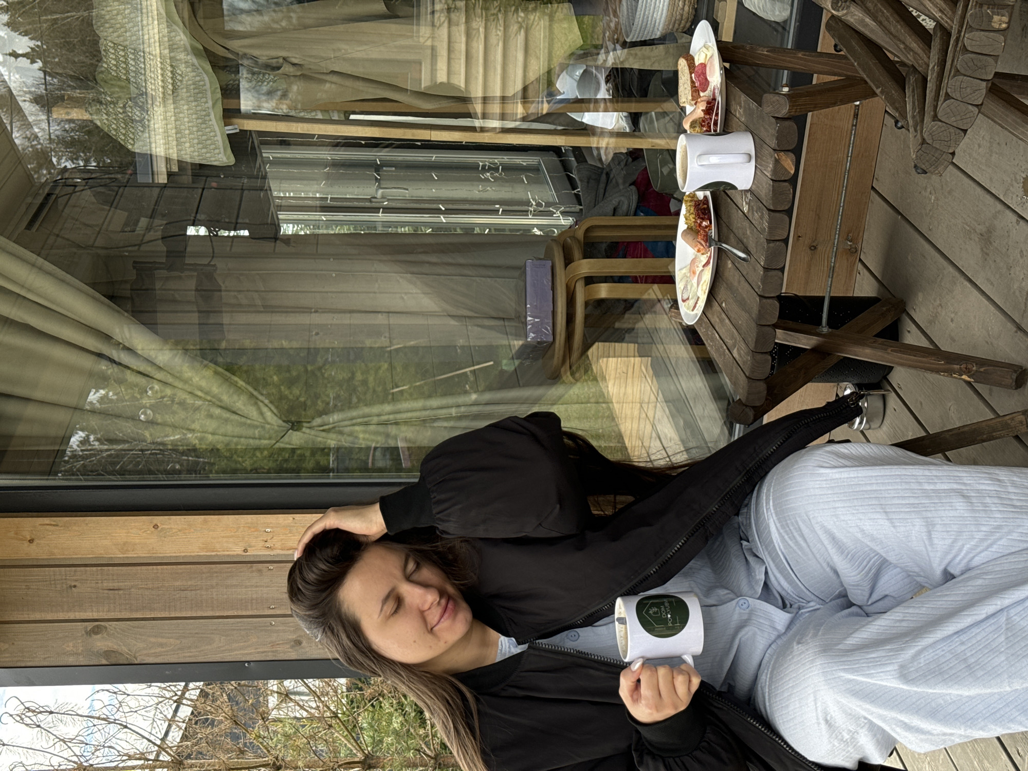 A woman with long brown hair and a black jacket is seated on a wooden structure, possibly a porch, in what appears to be the city of Литвиново, Russia. She is holding a white mug with green details. Next to her, visible through a large window, is a wooden table set for a meal with plates of food and another white mug. The reflection in the window shows the outdoor environment, including trees and possibly other buildings. The setting suggests a casual and relaxing atmosphere.