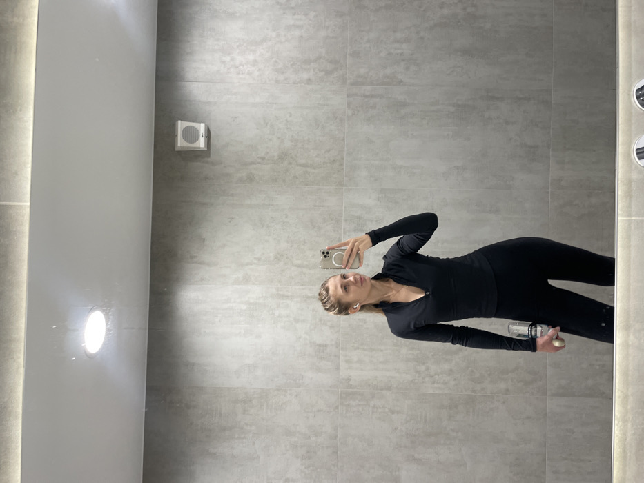 The image displays a woman taking a mirror selfie in what appears to be an indoor setting, possibly a gym changing room, bathroom, or a well-lit hallway. The image is oriented sideways, but when mentally rotated upright, the woman is standing, facing the mirror.
The woman has blonde hair, which appears to be pulled back, and is wearing a long-sleeved black athletic top with a partial zip or buttons at the neckline, paired with matching black leggings. She has small hoop earrings and a delicate necklace. In her right hand, she holds a smartphone with a clear case featuring a prominent metallic ring, likely for MagSafe, pointed towards the mirror to capture her reflection. In her left hand, she holds a metallic-colored bottle, possibly a water bottle or a shaker. Her expression is neutral as she looks towards her phone.
The background reflected in the mirror consists primarily of a wall covered in light grey, rectangular tiles with visible grout lines, giving a modern or industrial aesthetic. Mounted on this tiled wall, to the viewer's upper left of the woman, is a small, white square fixture that resembles a vent or a speaker. To the woman's left (viewer's right, if the image were upright), a light grey or white vertical panel or wall section is visible. This panel features a bright, vertical strip of light, likely an LED fixture, and a circular recessed light near its bottom. Part of her footwear, a black and white patterned shoe, possibly a checkerboard design, is partially visible at the bottom right edge of the reflection in the mirror. The lighting is bright and uniform throughout the scene. There is no visible text in the image.