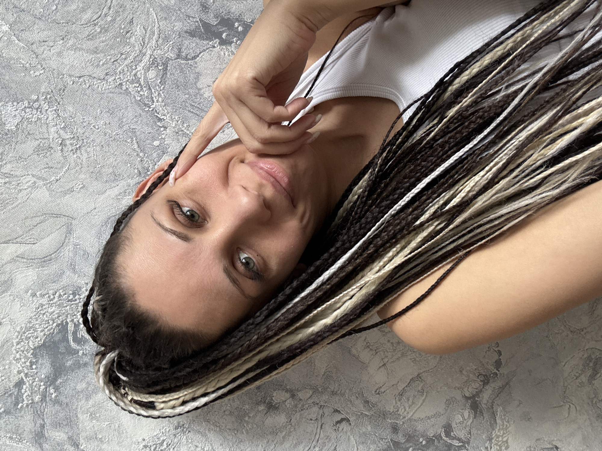 A woman lies on a gray textured surface. She is wearing a white tank top and has long braids that are a mix of brown, black, and blonde colors. Her right hand is touching her face near her right ear. The location of the photo is unknown and there is no indication it was taken in Rusanovo, Russia.