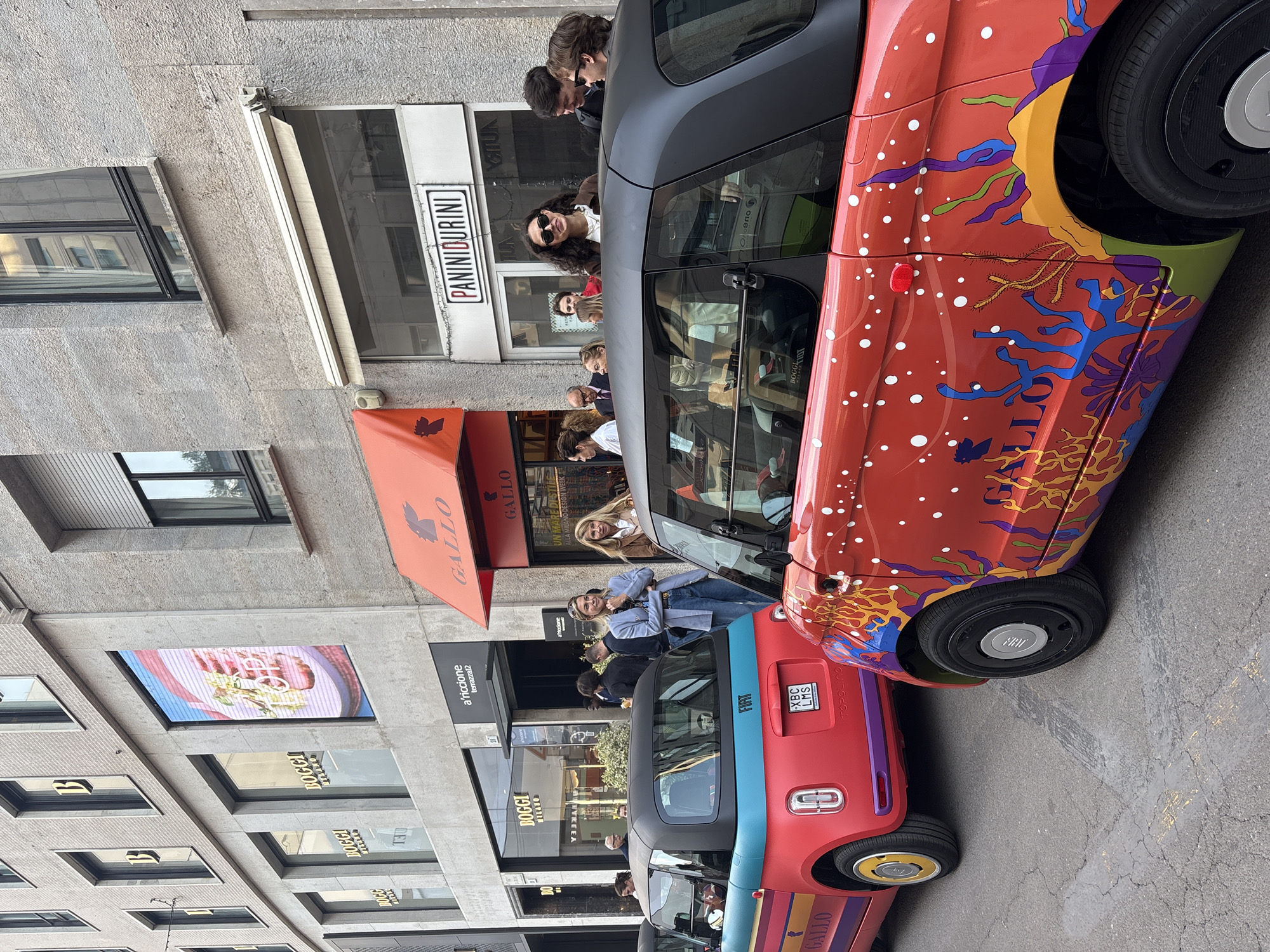 A vibrant city street scene in Milan, Italy, unfolds during what appears to be a clear day, likely related to Milan Design Week. In the foreground, two custom-painted Fiat Topolino electric cars are prominently displayed, drawing attention from passersby. The car in the foreground is predominantly red, adorned with an elaborate, colorful, and abstract marine-themed design featuring coral-like shapes in blue, purple, yellow, and green, accented with numerous white dots. The rear of a second Topolino is visible behind it, also red, with horizontal blue and yellow stripes and purple details, carrying a license plate "XBC LMS" and the text "TOPOLINO."
Several people are seen on the sidewalk, some observing the cars, others engaged in conversation or looking into shop windows. A woman with curly hair and sunglasses smiles towards the camera, as does another woman in a blue jacket.
The street is lined with buildings housing various establishments. Visible shop signs include "PANINIDURINI," "GALLO" (on red awnings), and "BOGGI MILANO" (on multiple storefronts). One window also shows "a'riccione terrazza12." A digital display embedded in a building facade showcases an image of food, possibly a steak or sandwich. A banner partially visible above one of the "GALLO" awn reads "UN MARE DI..." and clearly states "ALLA MILANO DESIGN WEEK," confirming the event context. The overall atmosphere appears lively and curious, with people appreciating the customized vehicles and the urban environment.