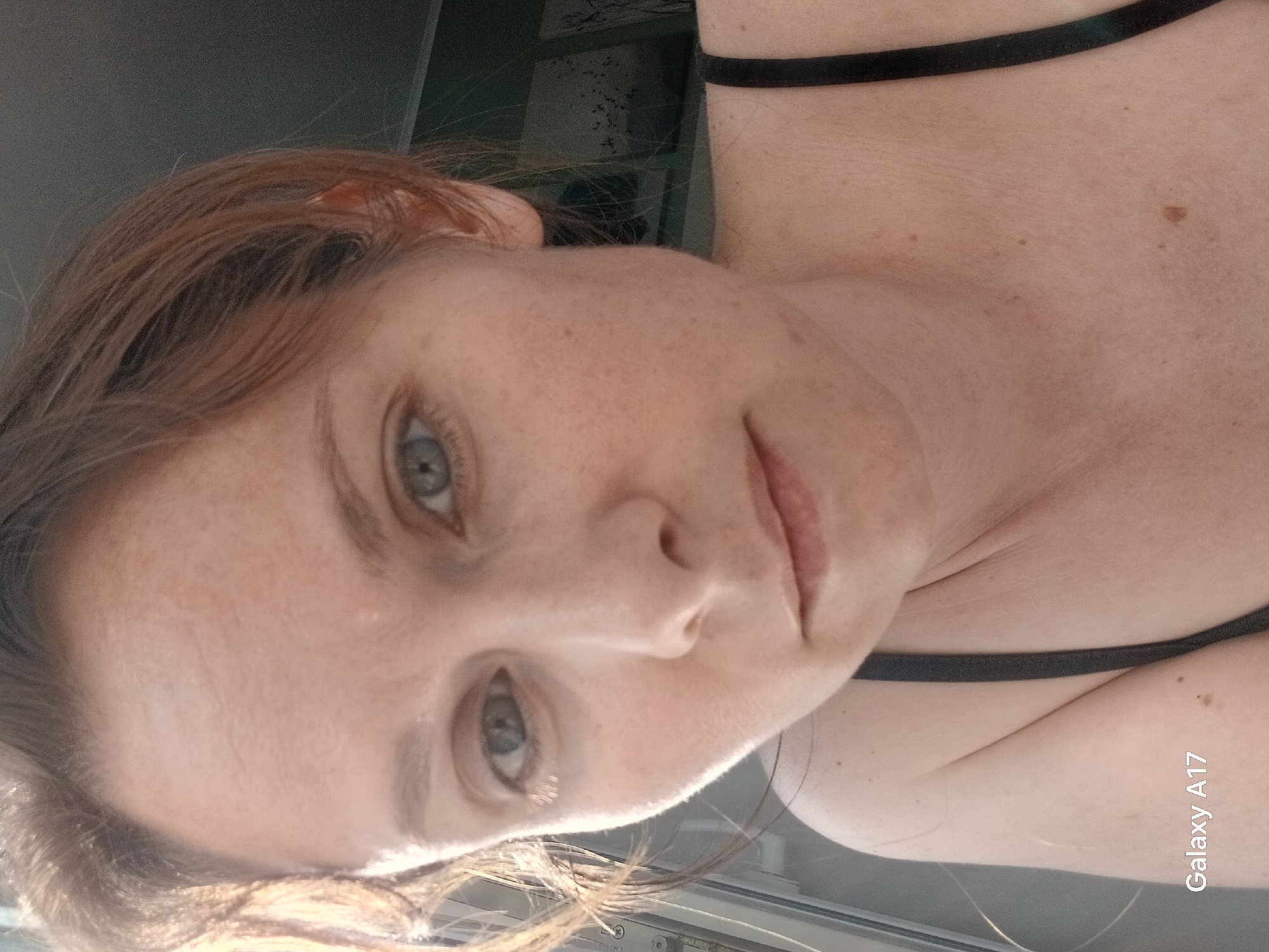 A fair-skinned woman with reddish-brown hair and grey eyes is shown in a close-up selfie. She is wearing a black top with thin straps. Her skin has visible freckles and a mole on her chest. The background is slightly out of focus, but there are hints of green and white that suggest a room or outdoor setting with plants or patterned wallpaper. There is a "Galaxy A17" watermark in the lower right corner of the image. The image does not provide sufficient information to determine a specific location in Yekaterinburg, Russia.