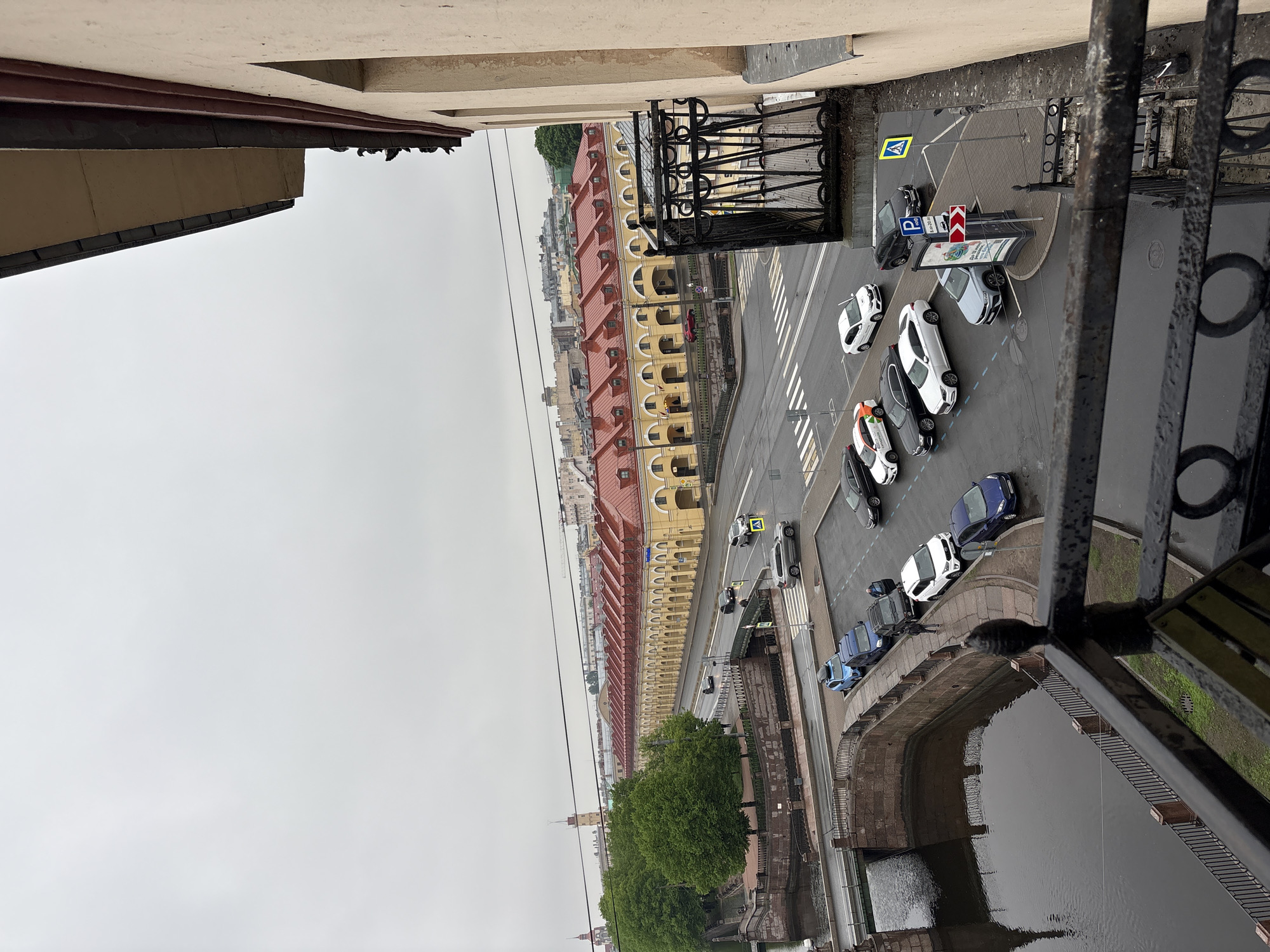A high-angle view from a building in Saint Petersburg, Russia, overlooks a street and canal on a cloudy day. The architecture includes yellow buildings with red roofs. Cars are parked along the street. A pedestrian crosswalk spans the street. Below, there is a canal with trees lining its banks. A bridge crosses the canal. Metal railings and part of the building from which the photo is taken are visible in the foreground.