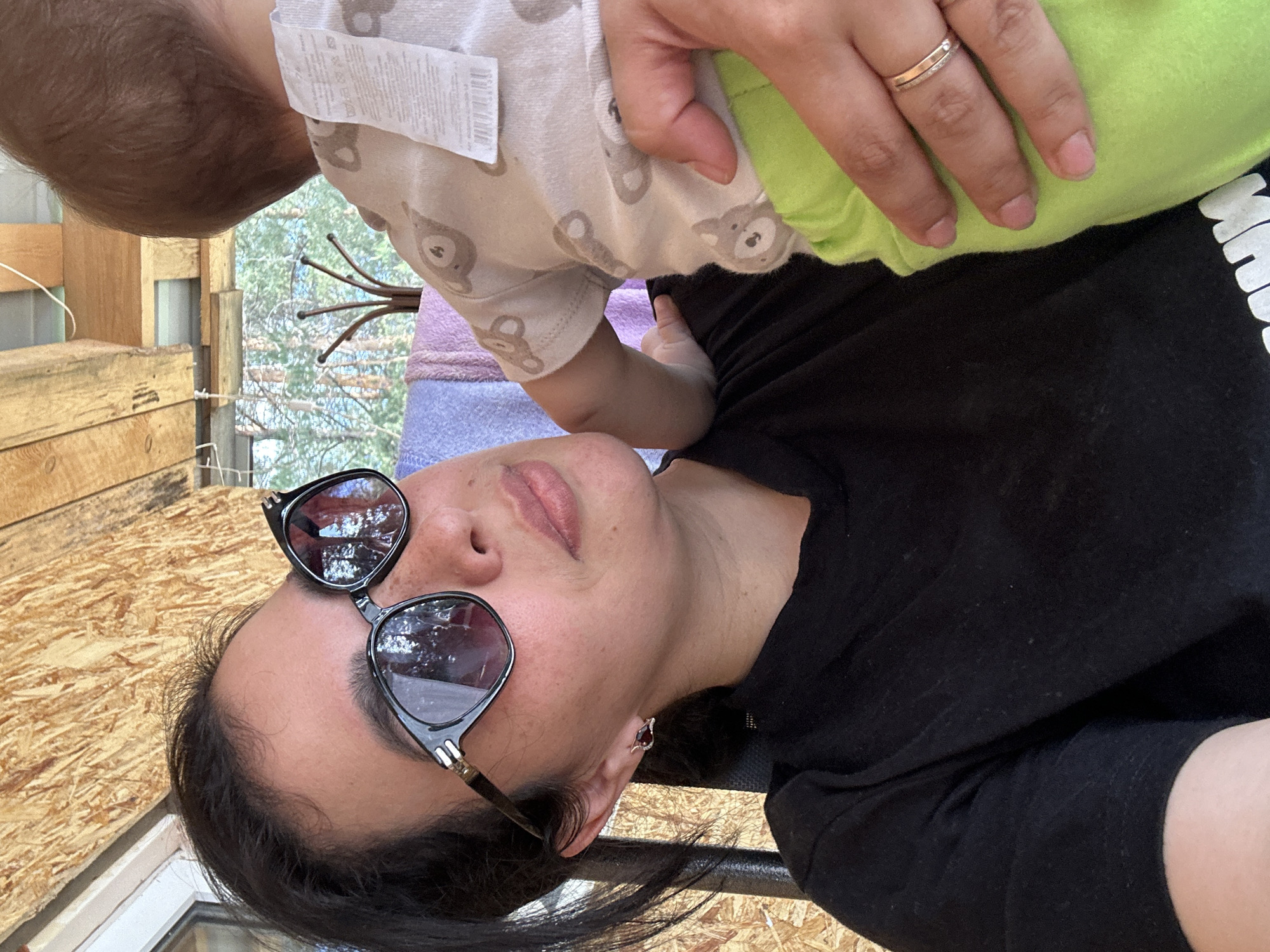 A woman wearing sunglasses and a black shirt is holding a baby. The baby is wearing a light-colored onesie with a teddy bear pattern and a lime green blanket. The woman is wearing a ring. The background shows an outdoor setting with wooden structures and foliage. The location is in the city of Unknown, Unknown.