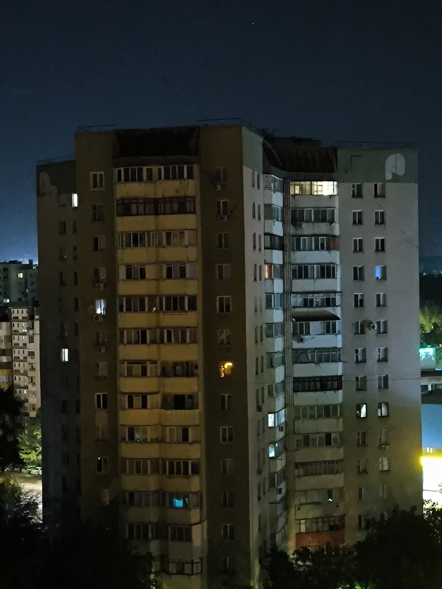 This is a nighttime, medium-distance, eye-level shot of a multi-story apartment building in Chișinău, Moldova. The building is tall and appears to be made of concrete, with numerous windows and balconies. Some windows are illuminated, suggesting occupancy. The building is situated in a densely populated area, with other similar buildings visible in the background. Trees and foliage are present in the foreground and around the base of the building, indicating a partially urbanized or suburban setting. The sky is dark, with a few visible stars. The overall impression is of a residential area at night.