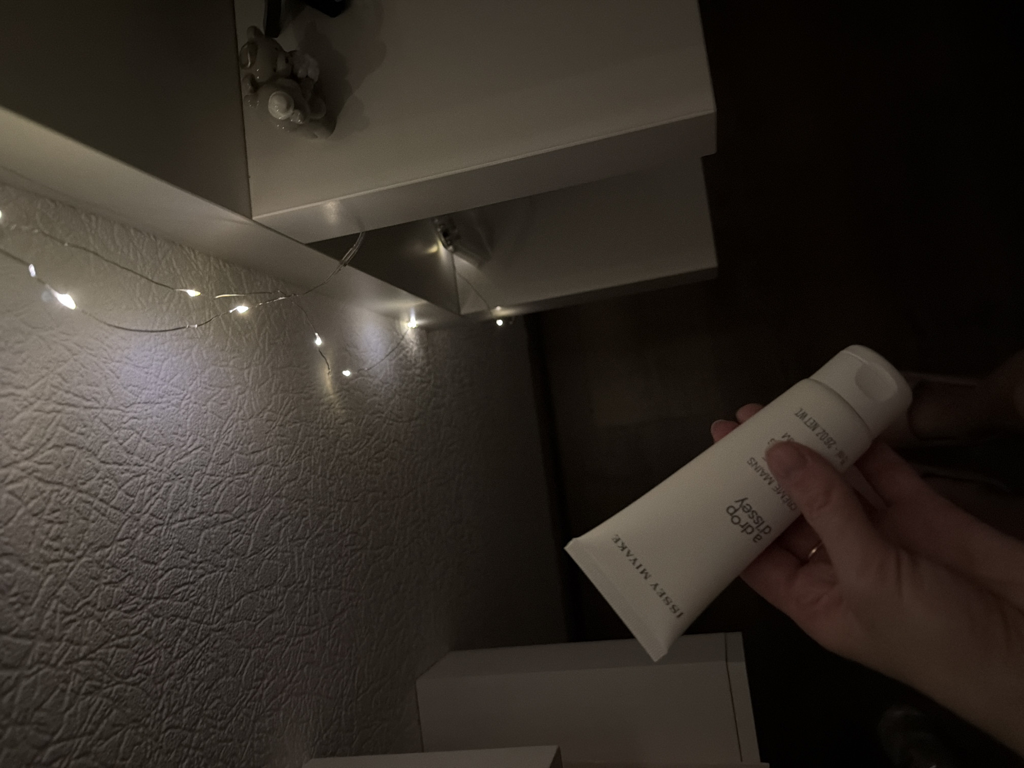 An overhead shot shows a person's hand holding a white tube of Issey Miyake "a drop d'issey" hand cream. The tube is partially obscured by the hand and is angled upwards. The background consists of a textured ceiling and white shelving units, with a string of fairy lights draped across the top. A small, beige figurine of a creature, possibly a bear or dog, sits on the shelf to the right. The lighting is dim, with the fairy lights providing small points of illumination. The scene appears to be indoors, possibly in a residential setting in Kyiv, Ukraine.