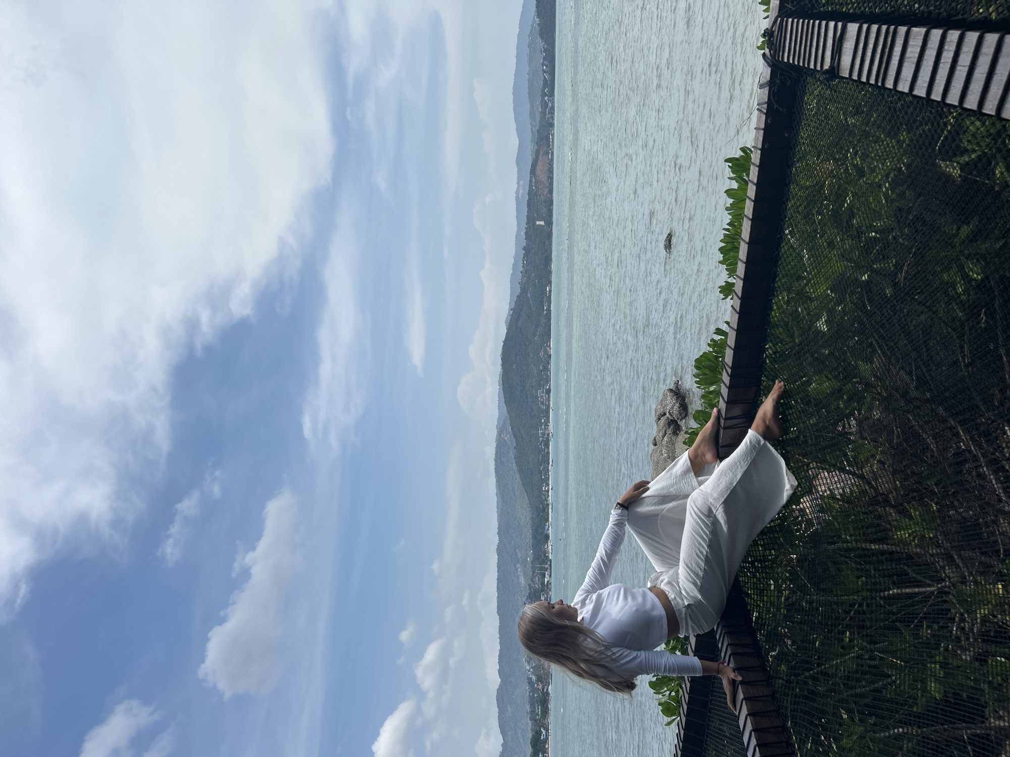 A woman sits on the edge of a large net structure, her legs extended over the netting and her arms resting on her lap. She is wearing a white long-sleeved crop top and white wide-leg pants. The woman is looking out towards the ocean and a lush, mountainous coastline. Boats are visible in the distance on the water. The sky is blue with scattered white clouds. The scene appears to be a scenic overlook in Ko Samui District, Thailand.