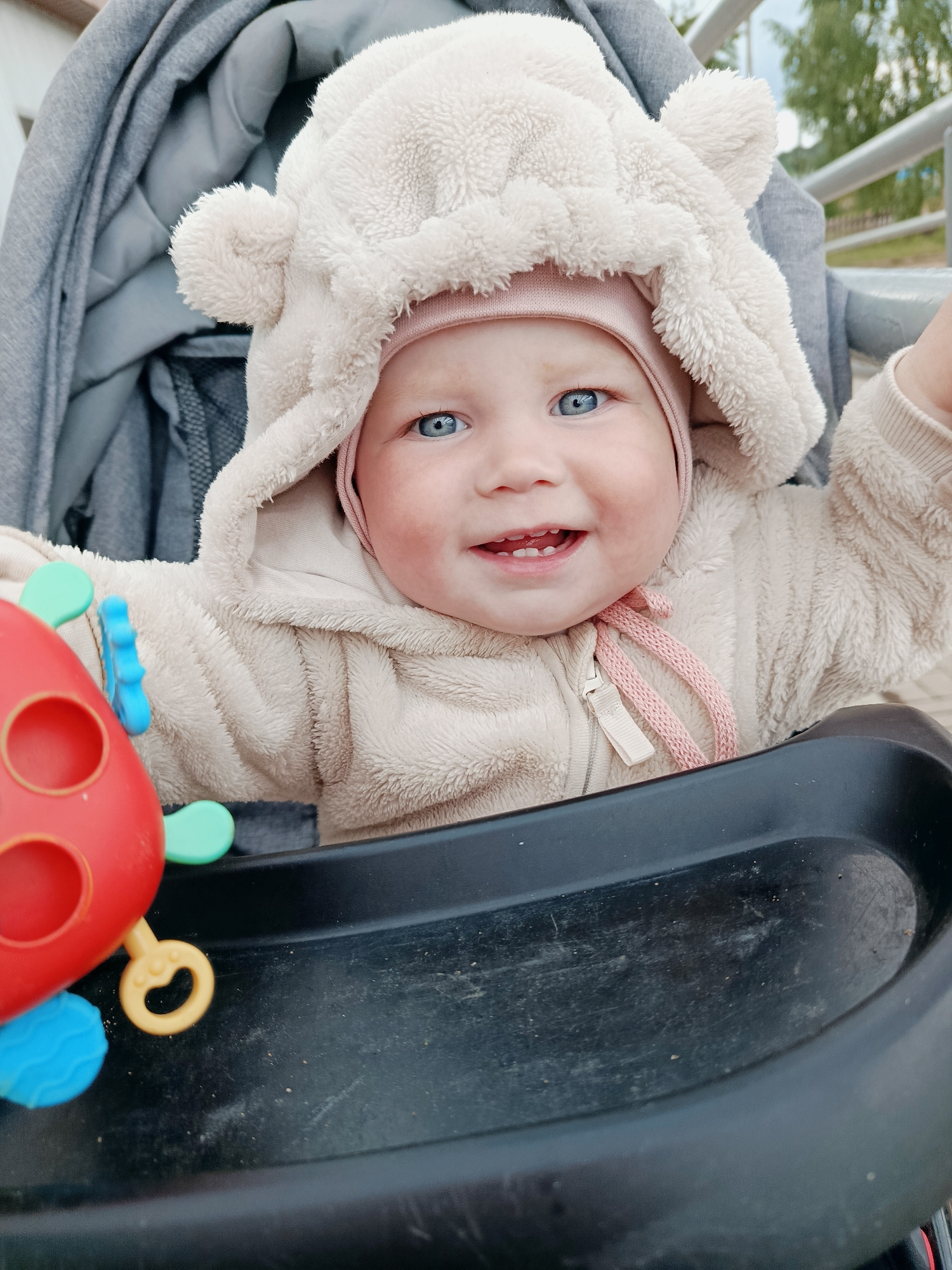 A baby with blue eyes is sitting in a stroller. The baby is wearing a light brown hooded fleece jacket with ears on the hood, and a pink cap underneath. The baby is smiling, showing a few small teeth. A red, yellow, and blue plastic toy is attached to the stroller's tray. The stroller is dark gray and black. In the background, there are glimpses of green trees and a gray fence, suggesting an outdoor setting. The location is in the city of Фоки, Russia.
