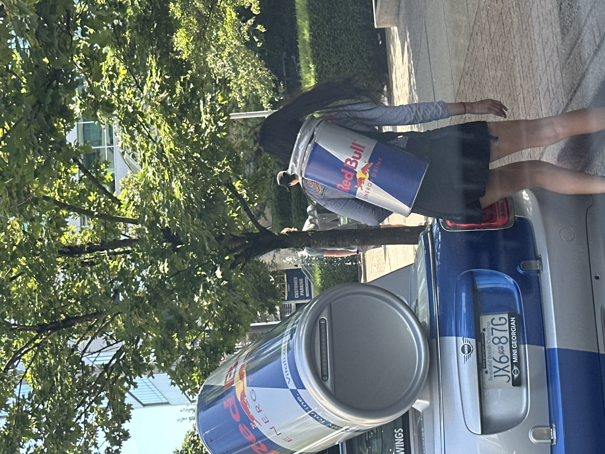 In Burnaby, Canada, a woman with long dark hair walks away from the camera, wearing a black skirt and a long-sleeved grey shirt. She is carrying a large, cylindrical Red Bull can on her back as a backpack. To her left, a silver and blue Mini Cooper is parked. On the trunk of the car is an even larger, similarly designed Red Bull can, resting on its side. The license plate of the Mini Cooper reads "JX6 87G" and indicates "Beautiful British Columbia" and "MINI GEORGIAN". In the background, trees with green leaves cast dappled shadows on a paved walkway. Beyond the trees, a modern building with a glass facade is visible, and further back, a sign reads "CUSTOMER PARKING". A man in a light-colored shirt and a man in a hat are also visible in the background, walking away from the camera. The overall scene appears to be an outdoor urban environment on a sunny day.