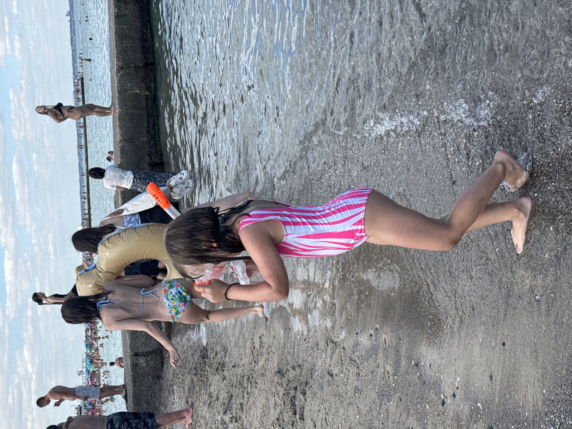 The image depicts a lively scene at a beach, likely in Odesa, Ukraine, given the context. Several people are present, enjoying the water and the shore. In the foreground, a young girl in a pink and white striped swimsuit is looking down at a snorkeling mask she is holding. She appears to be interacting with the water or preparing to use the mask. Next to her, another person is partially visible, wearing a floral bikini bottom.
In the background, a crowd of people is gathered, suggesting a busy beach day. Some are in the water, while others are on the shore. The sea is calm with gentle waves lapping at the pebbly beach. The sky is partly cloudy, indicating pleasant weather. The overall atmosphere seems relaxed and summery, with people enjoying a typical day at the seaside. A concrete structure runs along the water's edge, possibly a pier or a breakwater, which provides a vantage point for some individuals. There is no visible text in the image.