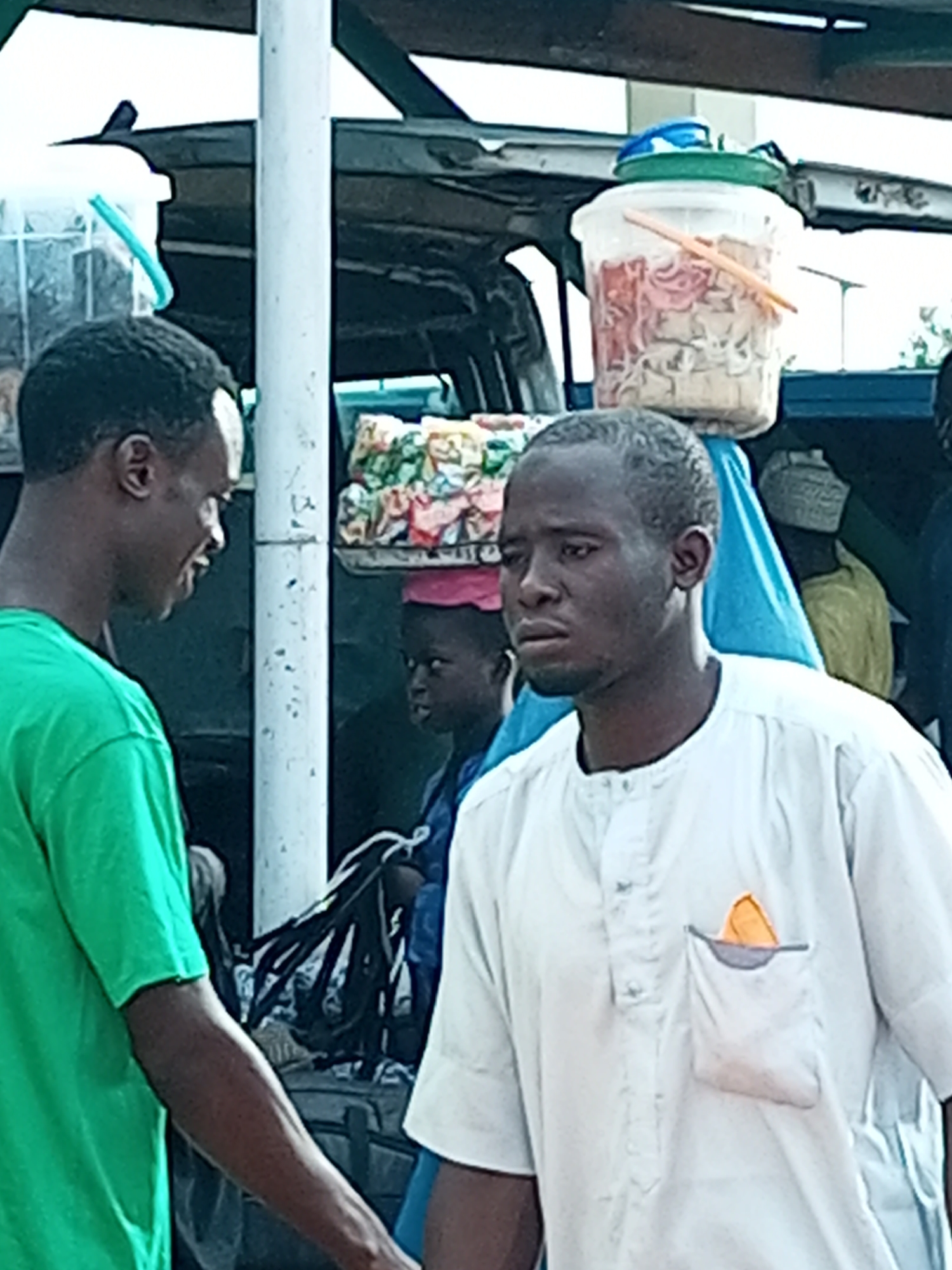 In Gombe, Nigeria, two men are standing near a roadside stall. The man on the left wears a green t-shirt and faces to the right, looking at the other man. The man on the right wears a white robe with an orange handkerchief peeking out of his chest pocket and faces slightly to the left, looking downwards. Behind them, a vehicle is parked, and various items are displayed on and around it, including large plastic containers filled with food items, bags of snacks, and other goods. A young person wearing a red head covering is visible in the background between the two men. The scene suggests a busy market or commercial area where vendors sell their wares.