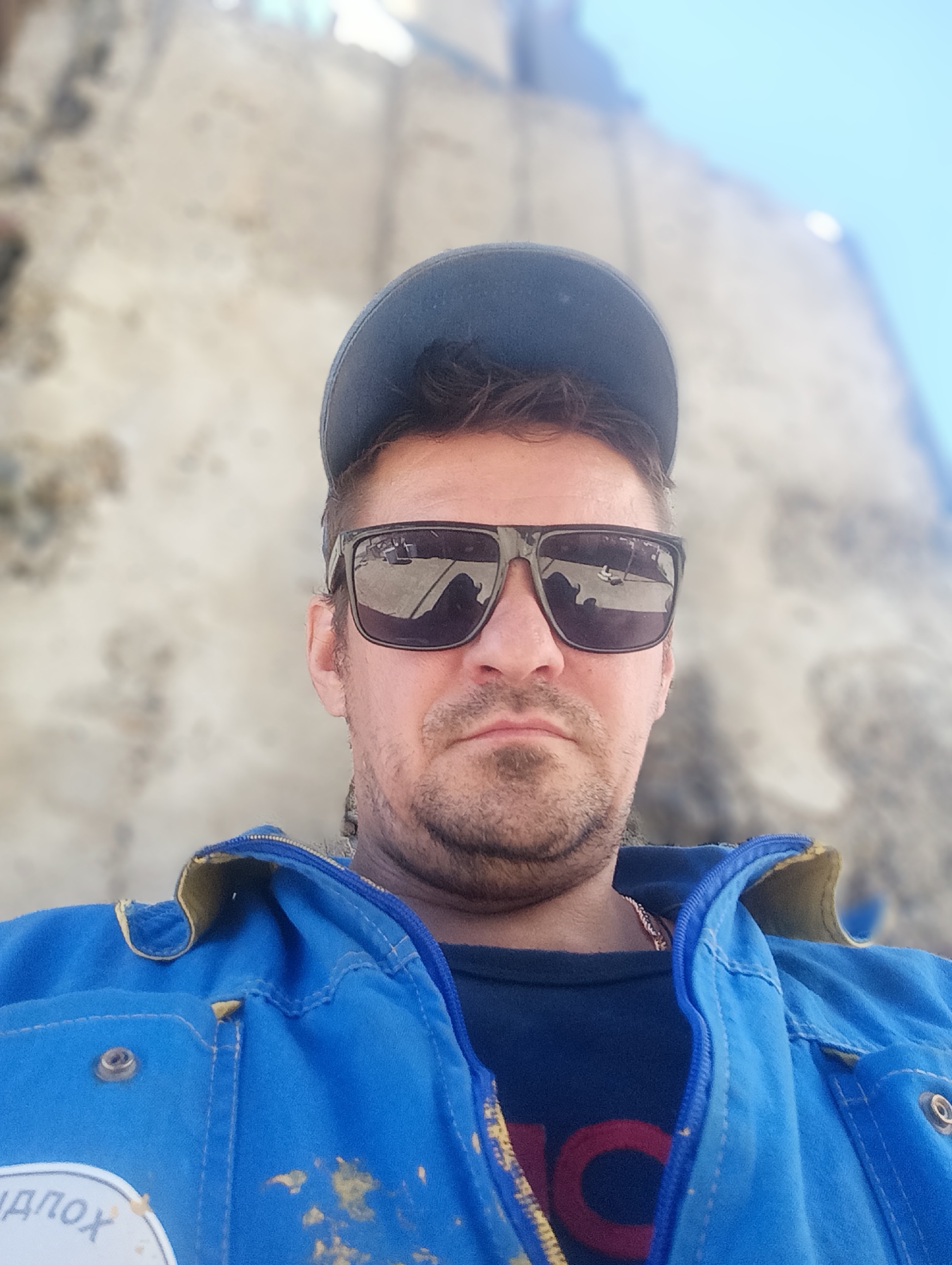 A man wearing sunglasses and a blue jacket is captured in a close-up, low-angle selfie. The man has a beard and is wearing a dark blue baseball cap. His blue jacket has yellow trim and appears to be slightly stained with a yellow substance. A portion of a logo with Cyrillic text is visible on the lower left of the jacket. The background is blurred, showing a rough, light-colored surface and a bright blue sky, suggesting an outdoor environment. The image is taken in daylight.