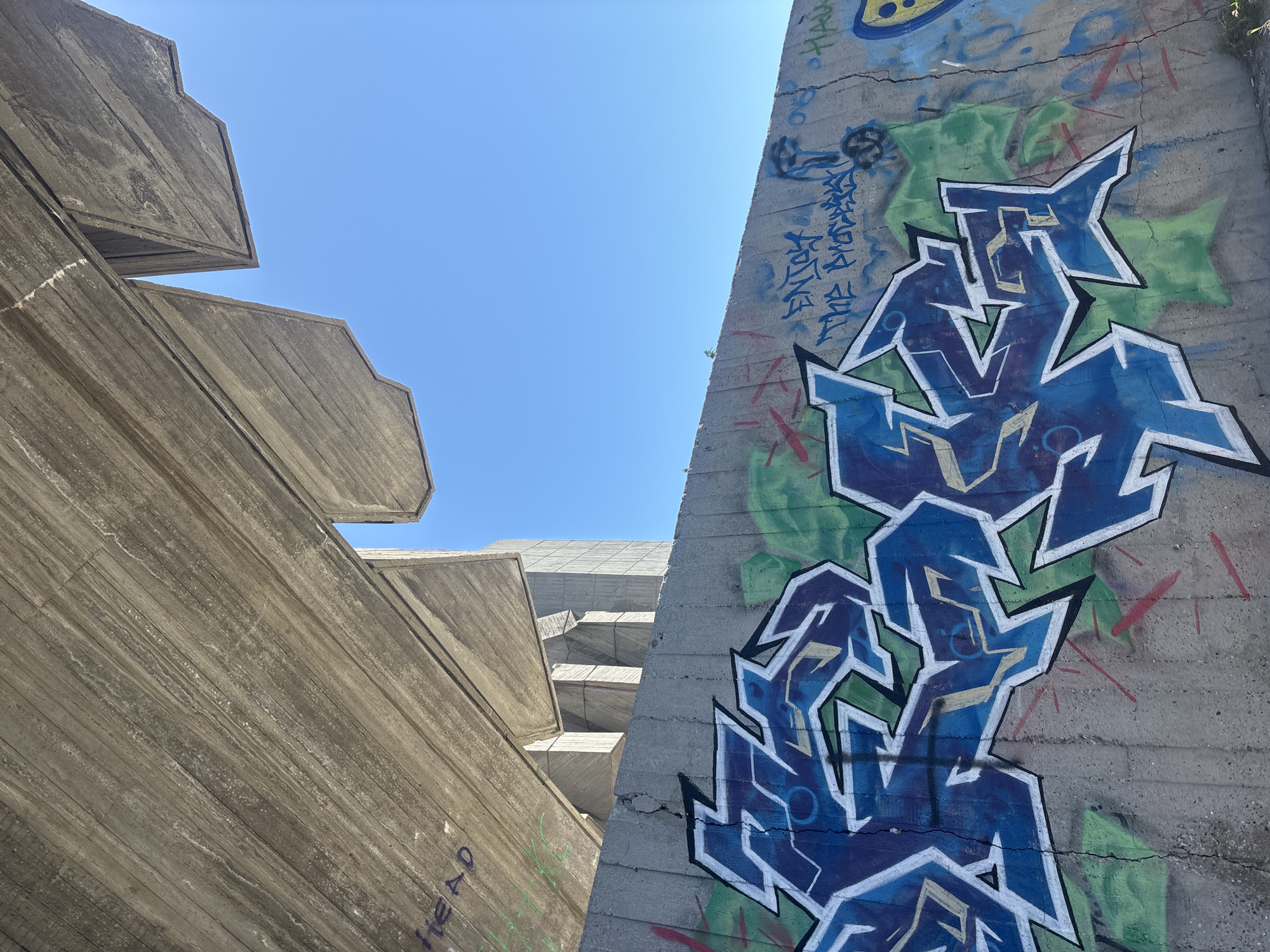 The photograph captures a low-angle view of modernist architecture in Varna, Bulgaria, under a bright blue sky. The left side of the frame features a section of a large, concrete building composed of stepped, cantilevered levels. The surface is textured and weathered, showing horizontal striations. Near the bottom left, faint graffiti spells "HEAD".
To the right, another concrete structure is visible, covered in vibrant graffiti art. The main piece is a stylized design in shades of blue and green with white outlines. Additional scribbles and tags are scattered around the larger artwork. The concrete surfaces of both structures appear aged and worn. The scene lacks any human presence.