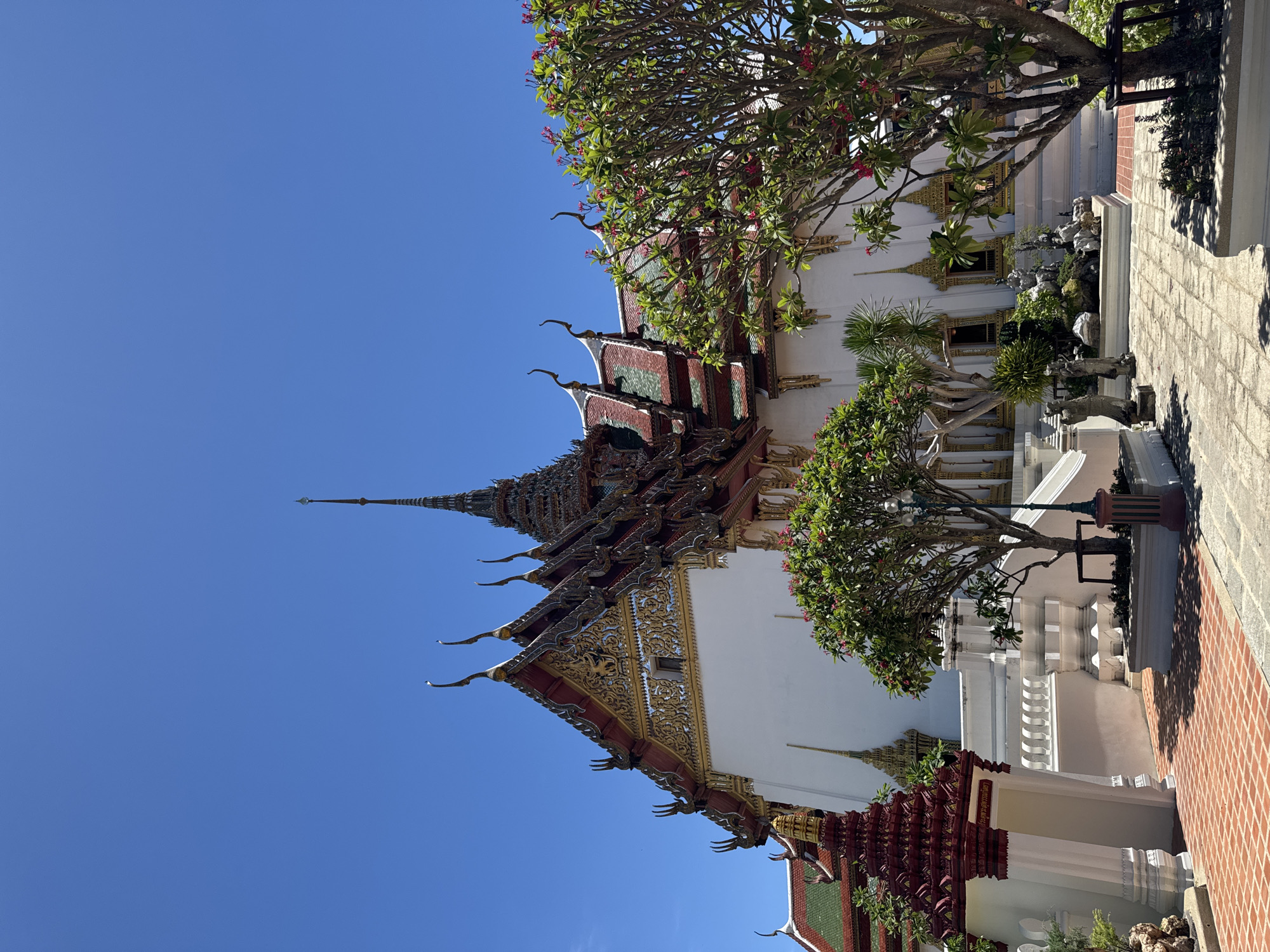 The image shows a building in Bang Pu Subdistrict Municipality, Thailand, under a clear blue sky. The building has a white facade with ornate gold details and a multi-tiered roof with red, green, and gold accents. Decorative pointed finials extend from the roof edges. A tall, tapering spire rises from the top of the roof. Trees with green leaves and red flowers are positioned near the building. A portion of a brick-paved area is visible in the foreground. The overall composition suggests a traditional Thai architectural style.