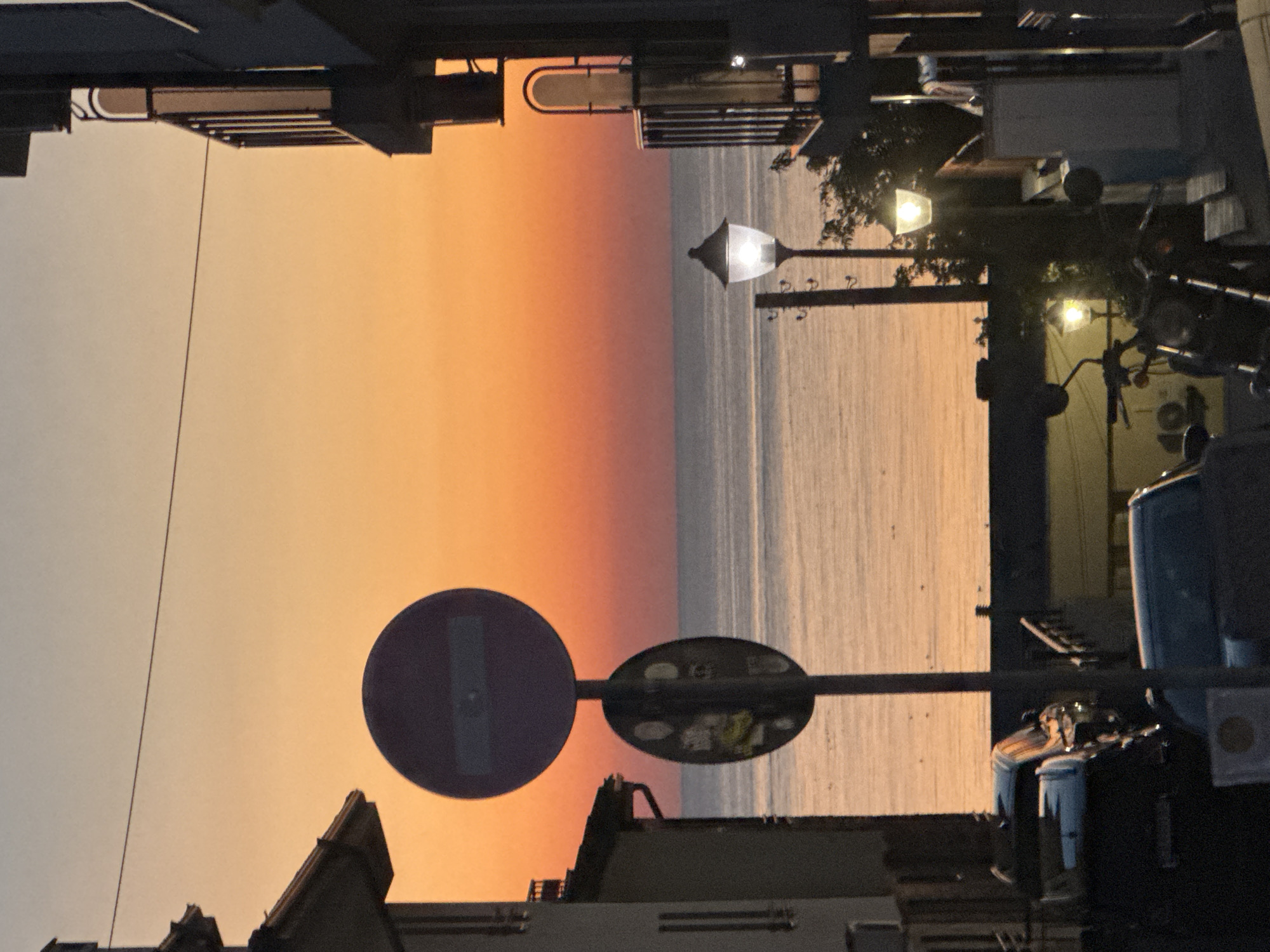 The image depicts a street scene at sunset in Sozopol, Bulgaria. A large traffic sign, a red circle with a horizontal white bar, stands prominently in the foreground, obscuring a portion of the vibrant orange and pink sky. Below it, a second, smaller circular sign is visible, featuring lighter colored circles. The sea is visible in the background, reflecting the warm hues of the sunset. On the right side of the image, a building with balconies and a street lamp illuminates the foreground. On the left, another building is partially visible. Several vehicles, including cars and a motorcycle, are parked or present on the street. The overall atmosphere suggests an evening setting, likely in a coastal urban area.