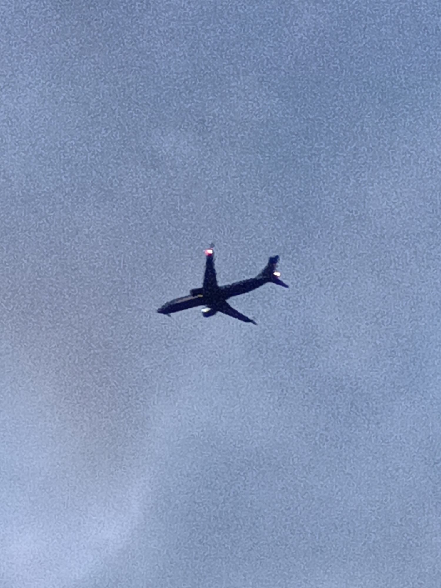 The image shows an airplane flying in the sky. The airplane is dark in color and has two engines visible. There are red lights on the tail and a white light on the underside of the wing. The sky is a muted blue-gray with some grainy texture. There is no indication of the location being Prague, Czechia.