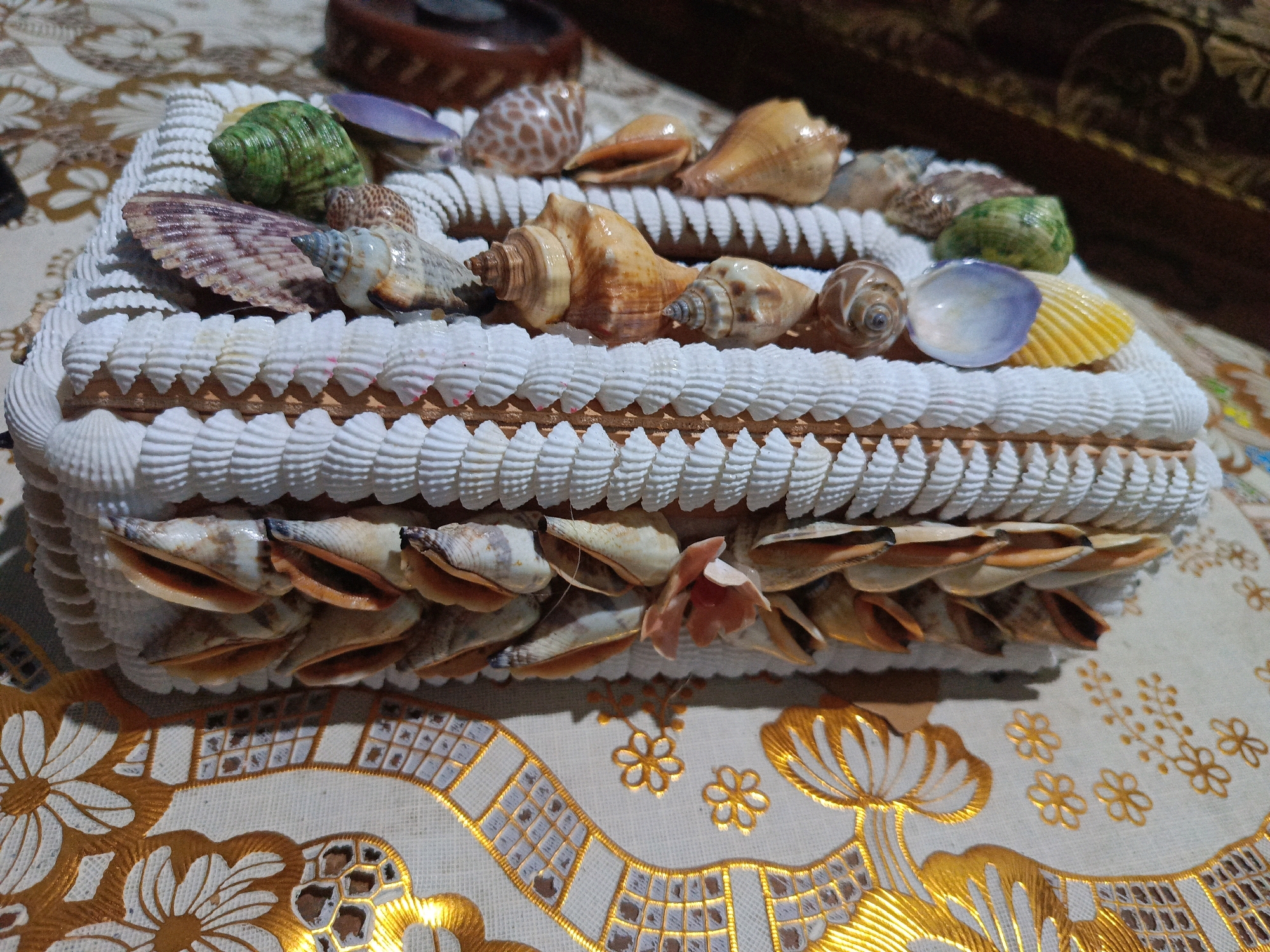 A close-up, high-angle shot captures a decorative tissue box adorned with various seashells. The rectangular tissue box is covered on its sides and top with white, textured shells, resembling ropes and rows of smaller shells. The top of the box is further embellished with larger, more prominent seashells, including a green spiral shell, a purple bivalve, and several brown and tan conch shells. The bottom half of the box features a dense arrangement of elongated, pointed shells facing outwards.
The tissue box rests on a fabric surface with a pattern of gold embroidery. The embroidery depicts stylized flowers and geometric shapes, creating a rich and intricate border along the lower portion of the image. The background shows a darker, out-of-focus area, possibly a wooden surface or furniture. The lighting is dim, casting shadows that add depth to the scene. The image appears to be taken indoors, likely in a home environment in Kalang, Indonesia, judging by the decorative style and the mention of the location. There are no people or explicit actions depicted; the focus is solely on the handcrafted tissue box and its surroundings.