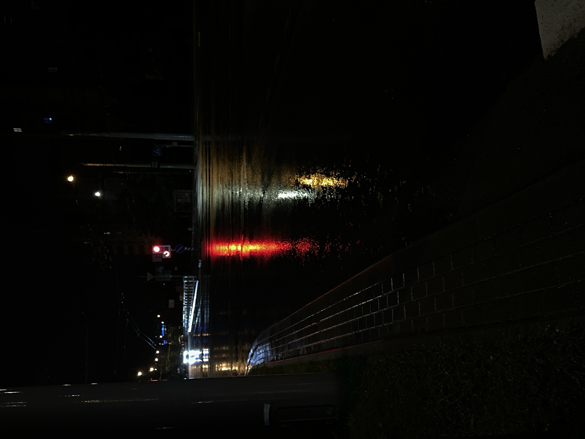 The image depicts a nighttime street scene in Minsk, Belarus, likely after rainfall, given the wet asphalt reflecting lights. A traffic light is visible, showing a red signal. A car with illuminated headlights is in motion on the street. The sidewalk, made of brick pavers, is also wet. Streetlights and building lights are scattered throughout the background, adding to the nocturnal ambiance. The overall atmosphere is dark and slightly moody, typical of a city at night with wet surfaces.