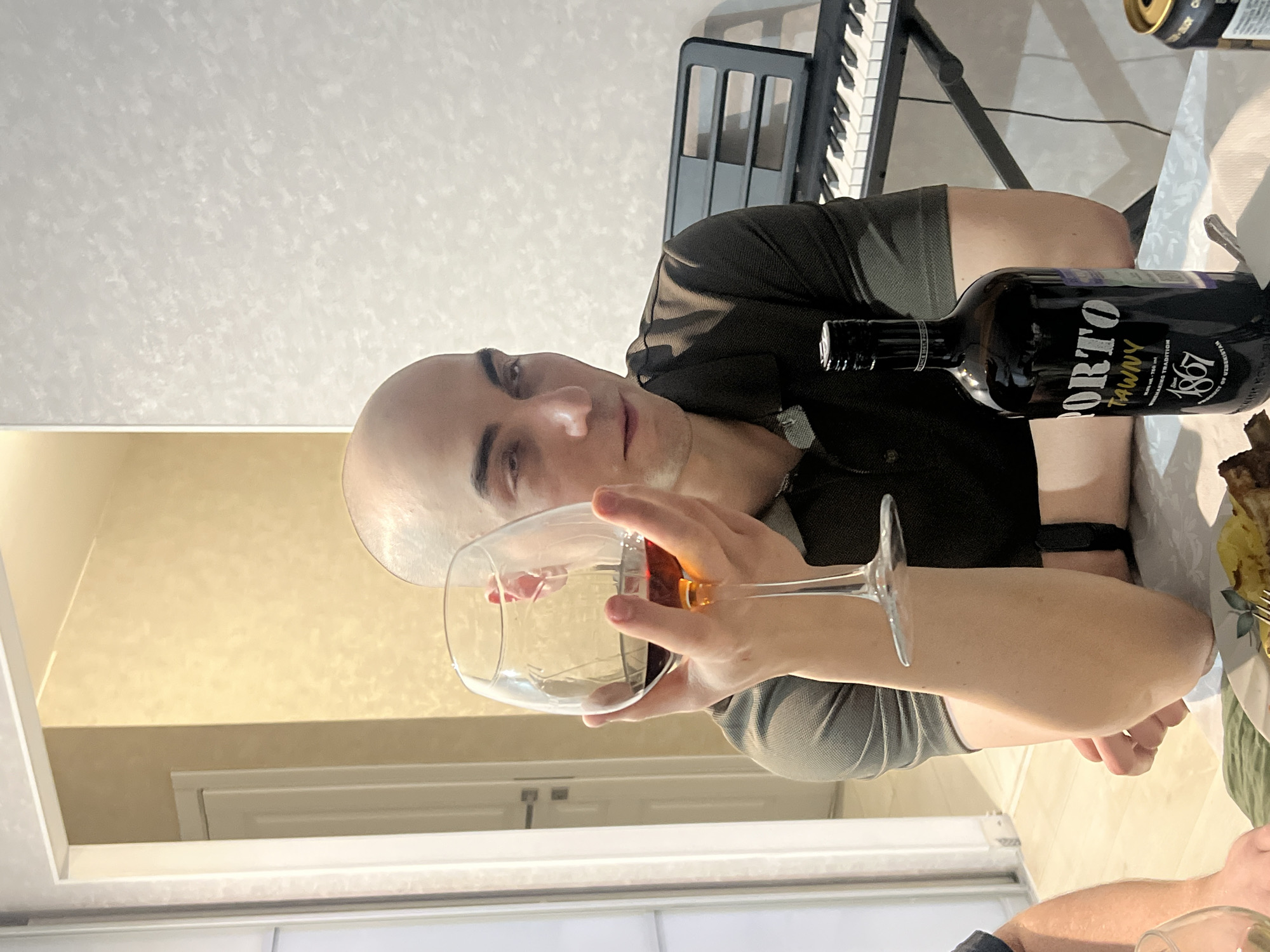 A man with a shaved head is holding a glass of tawny-colored liquid, likely wine, in his right hand. He is sitting at a table with a bottle of Porto Tawny 1867 wine nearby. A portion of a keyboard is visible in the background. The location appears to be indoors, possibly a residence. The city and country are Chaikovsky, Russia. Another person's arms and the edge of their glass is visible at the bottom of the image.
