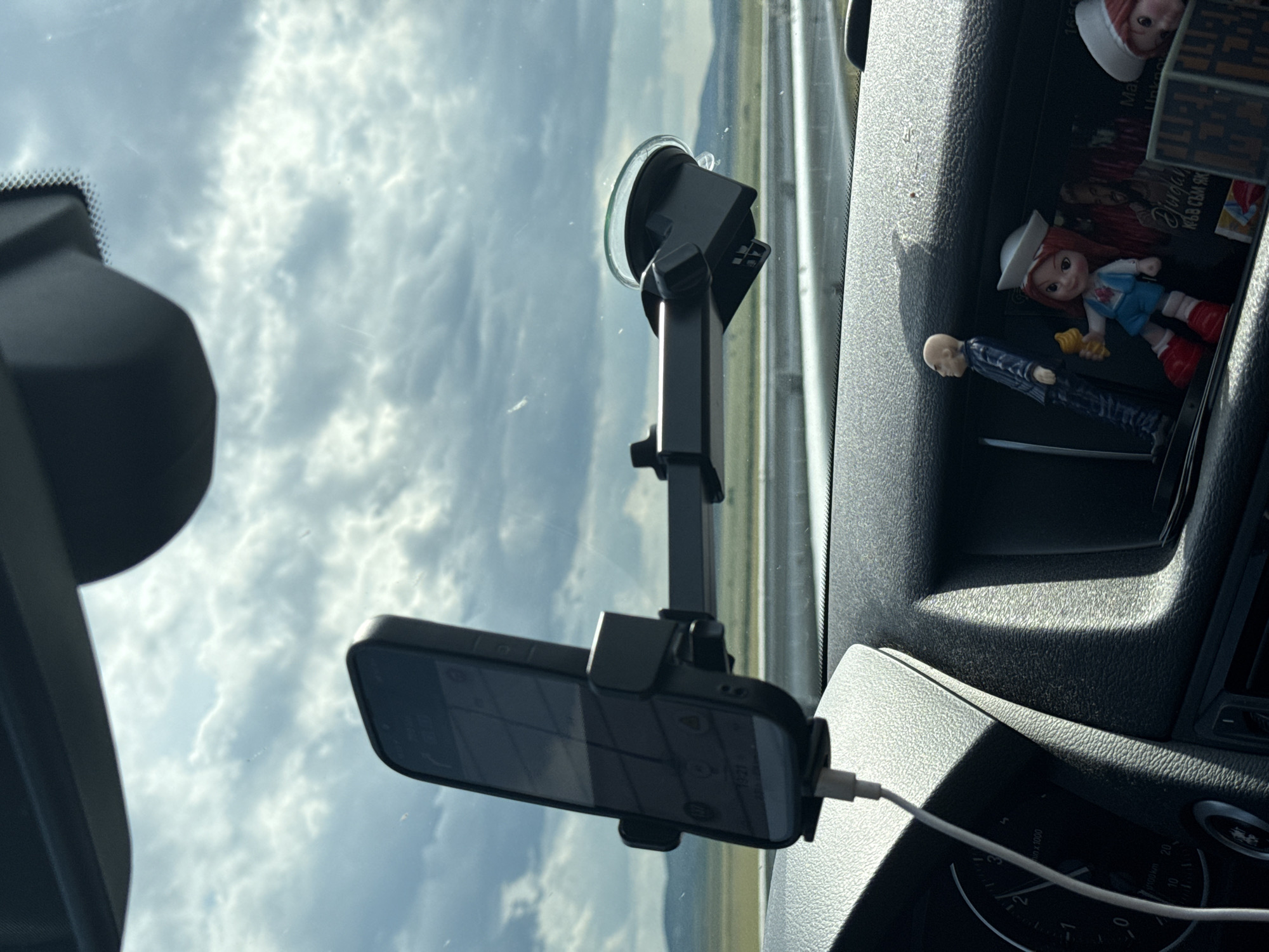 A smartphone is mounted on a windshield by a suction cup mount, with the phone displaying a navigation app. The car's dashboard is visible below the mount, featuring a charging cable plugged into the phone. Several small figurines, including a bald man in a suit and a girl in a sailor outfit holding an ice cream cone, are placed on the dashboard. The car is on a road, indicated by the presence of a highway barrier and a landscape of fields and hills under a cloudy sky. The scene is set in the city of Polsko Padarevo, Bulgaria.