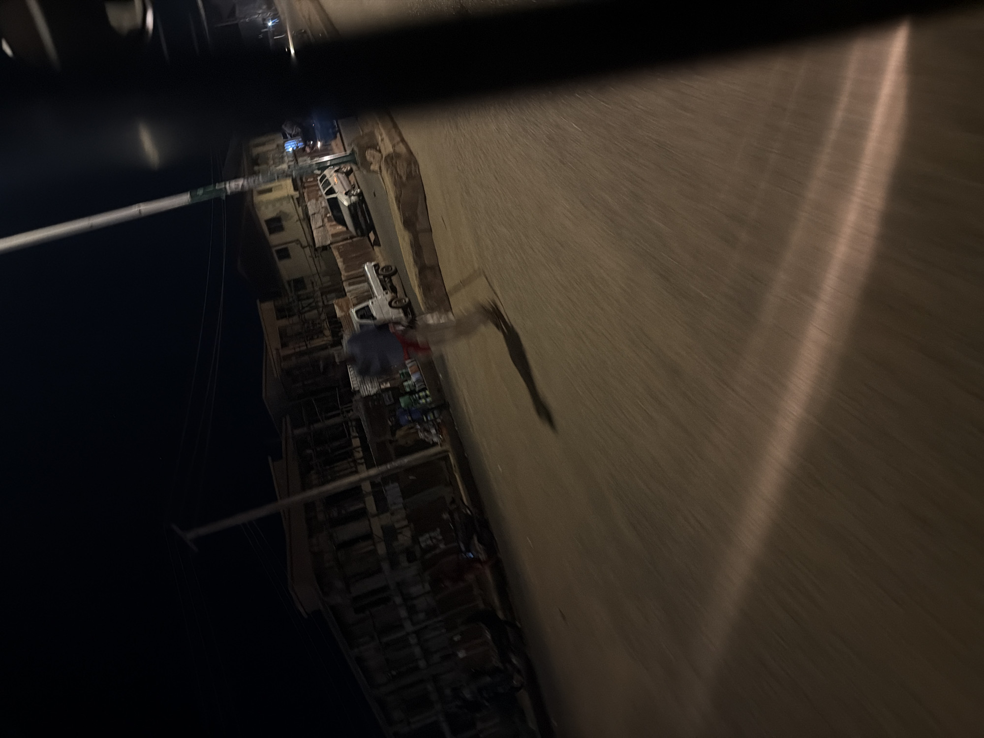 The image, taken at night in Bukuru, Nigeria, captures a street scene with low lighting and motion blur, suggesting movement from a vehicle. The primary subjects are two white pickup trucks and a person walking. One truck is parked to the left, and another is slightly further down the road. A person in a dark top and light-colored pants is walking on the right side of the road, casting a shadow.
Behind the parked truck on the left, there are buildings with multiple floors, appearing to be residential or commercial structures. The architecture suggests a dense urban or semi-urban environment. The overall scene is dark, with artificial light sources illuminating parts of the road and buildings. The time of day is clearly nighttime.
The motion blur in the image, particularly on the road surface which shows streaks of light, indicates that the photo was likely taken from a moving vehicle. This creates a sense of a fleeting glimpse of the street as it passes by. The weather appears to be clear, as there are no visible signs of rain or fog. The location context of Bukuru, Nigeria, provides a specific geographical setting for this nocturnal street view. There is no visible text in the image.