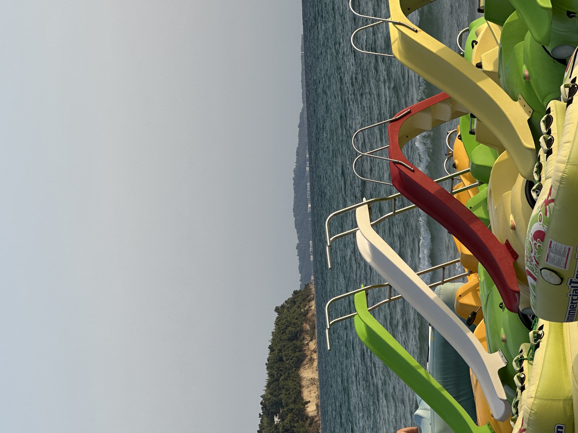 In Kassandra Municipal Unit, Greece, a cluster of brightly colored water slides, predominantly green, white, red, and yellow, are arranged on a sandy beach, facing a calm, teal-colored sea. The water is slightly choppy, with small waves lapping the shore. In the background, a verdant, tree-covered hillside slopes down to the water, with a few buildings visible on its crest. Further in the distance, another, lower-lying coastal area with structures can be seen. The sky is a pale, hazy blue, suggesting a late afternoon or early morning light. The slides appear to be part of a water park or rental service, likely intended for use in the sea.
