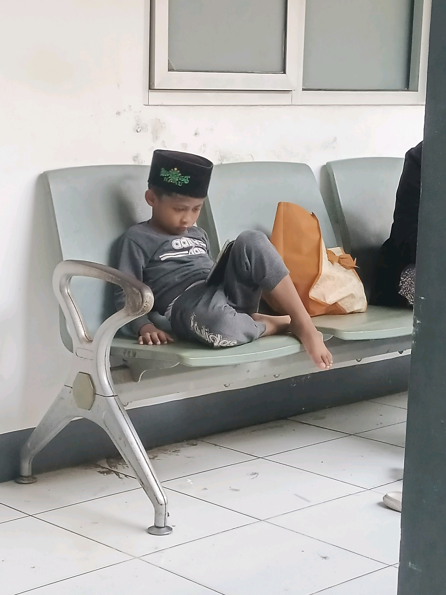 A young boy wearing a black skullcap is sitting on a grey plastic bench in what appears to be a waiting area. He is wearing a grey long-sleeved shirt and grey pants. He is looking down, seemingly at a book or device in his lap. To his right, there is a brown paper bag and next to it, a person in dark clothing, partially visible. The bench is made of grey plastic with metallic legs and is situated in a room with white walls and a tiled floor. The overall environment suggests a public or institutional setting, possibly a clinic or a government office in Kasturi, Indonesia.