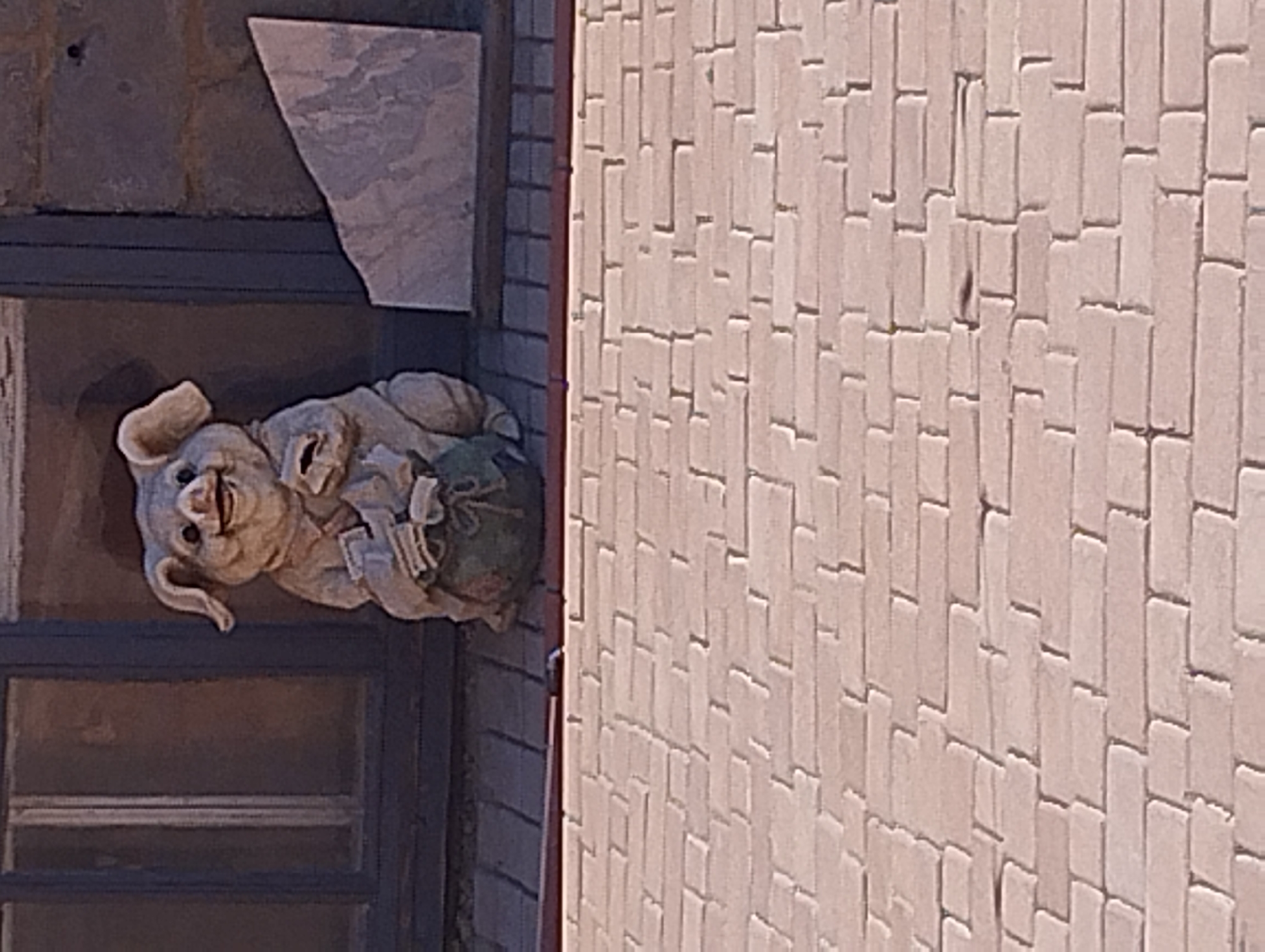 A ceramic statue of a pig is positioned in front of a window. The pig statue is holding a sack in its arms, and its face is smiling. The pig statue is on a ledge, with a brick wall in the foreground and a building in the background. The statue is made of a light brown material and is about knee-high. The window is made of dark wood and has a reflection of the pig statue in it. The brick wall is made of red bricks and is laid in a stretcher bond pattern. The building is made of stone and has a pitched roof.