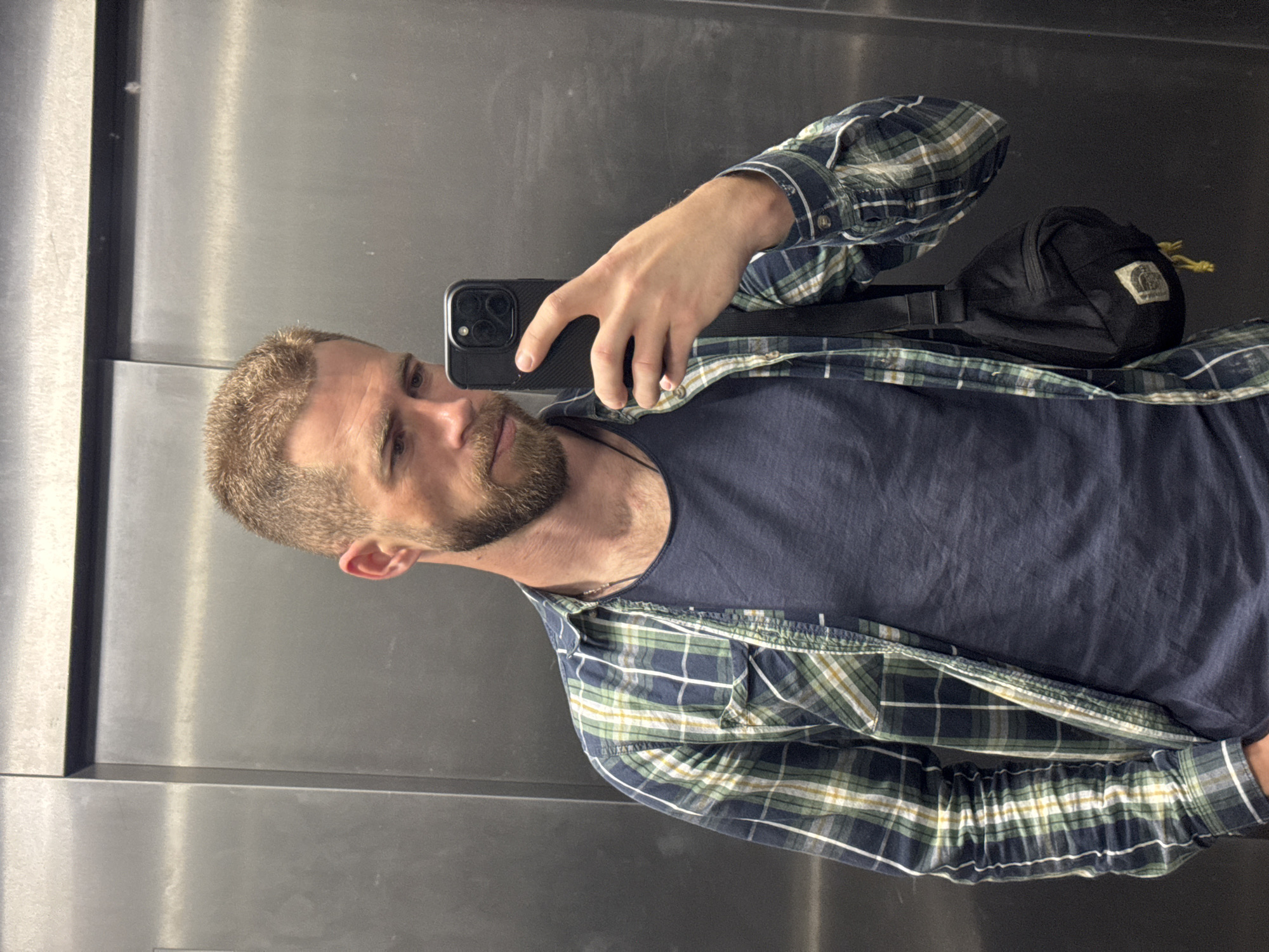 A man with short, light brown hair and a beard takes a mirror selfie in what appears to be an elevator or a space with brushed metallic walls, set in Budva, Montenegro. He is positioned facing slightly to his left, holding a dark-colored smartphone with a visible multi-lens camera bump in his right hand to capture his reflection. His expression is neutral to contemplative.
He is dressed casually in a dark blue t-shirt, over which he wears an unbuttoned long-sleeve plaid shirt featuring a pattern of dark blue, green, and white checks. A thin silver chain is visible around his neck. A black fanny pack or sling bag is worn across his shoulder, featuring a faded, light-colored patch with a mountain-like logo on its front, and a yellow drawstring or cord extending from it. The metallic walls behind him show some subtle reflections and minor surface imperfections, characteristic of a public indoor setting. No specific time of day or weather conditions are discernible. There is no clearly legible text visible on clothing, the bag (apart from the indistinct logo), or the background.