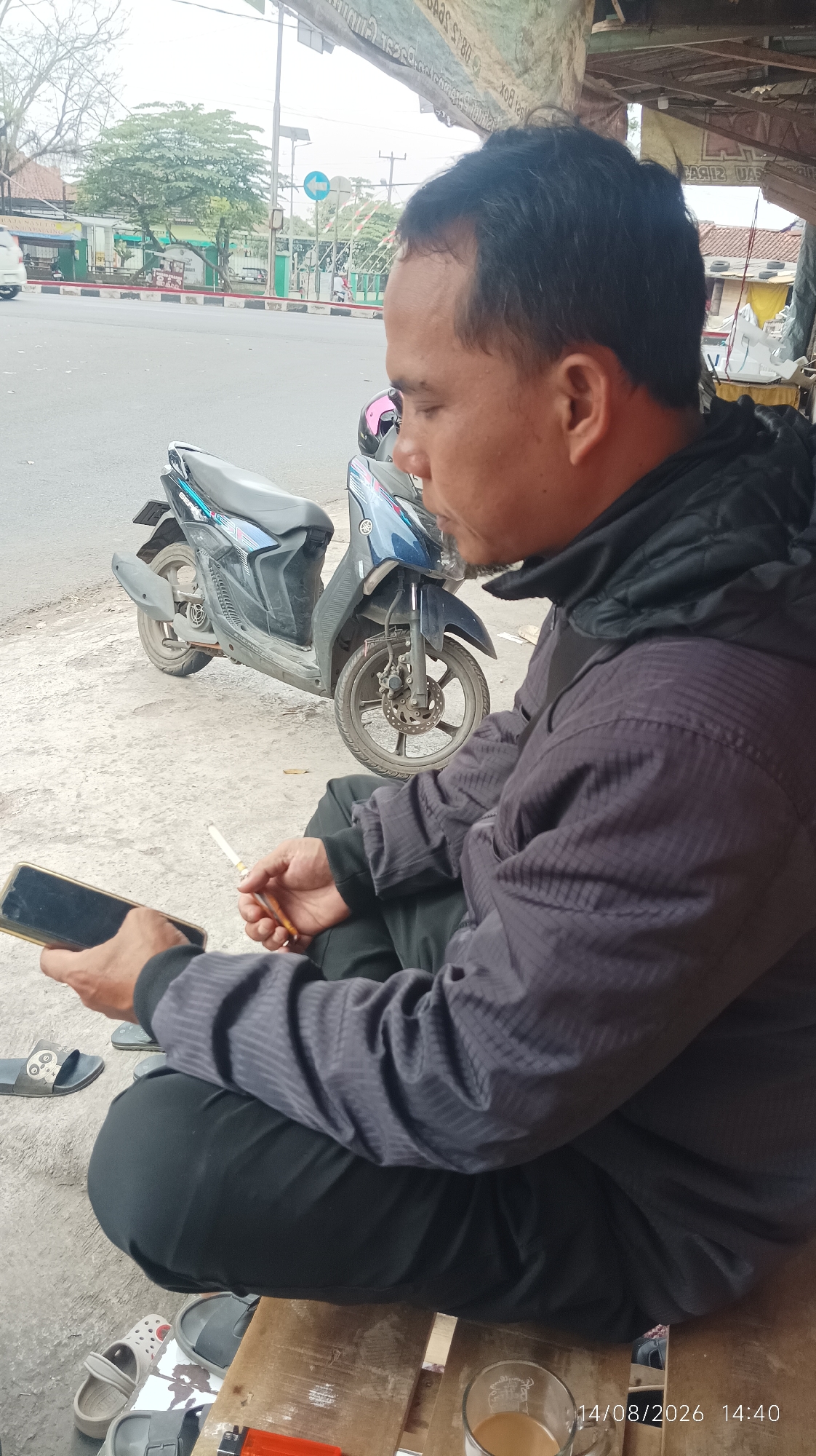 A man is sitting on the ground, holding a cigarette and a smartphone. He is wearing a dark jacket and dark pants. A blue and black motorcycle is parked behind him. The scene appears to be on a street in Tasikmalaya, Indonesia, with a roadside structure and a busy street in the background. There are also several pairs of sandals on the ground near the man. A cup of coffee is on a wooden surface in front of him. The timestamp indicates the photo was taken on August 14, 2026, at 2:40 PM.