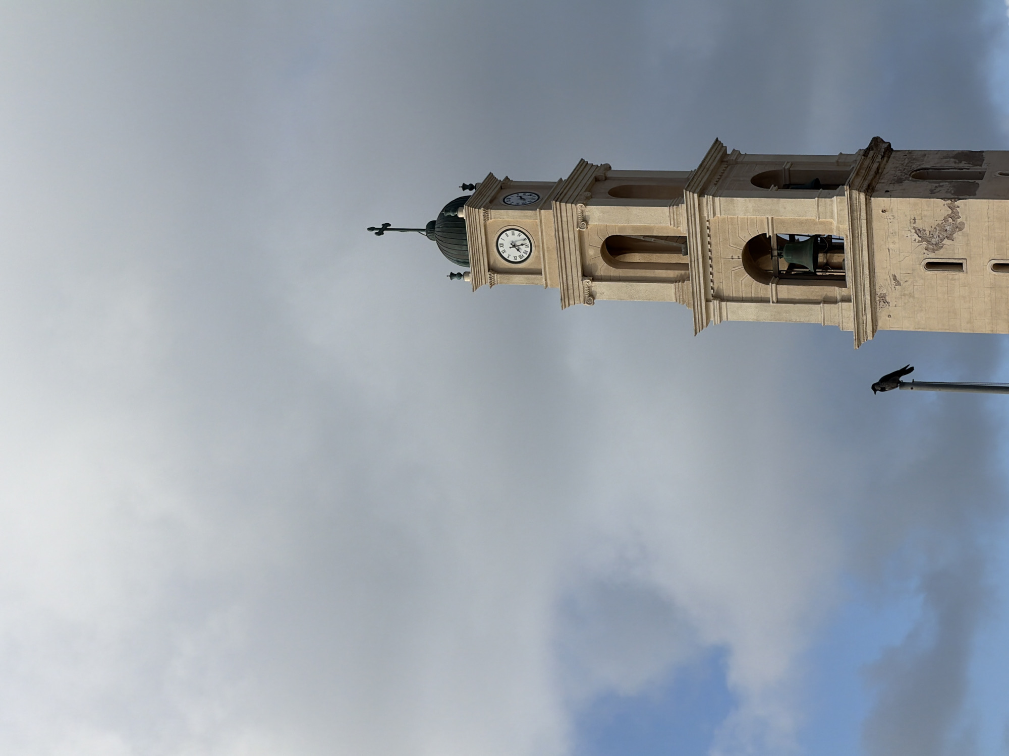 The image is a vertically oriented, close-up shot of St. Peter's Church in the city of Tel Aviv, Israel. The church tower, made of light-colored stone, is prominently featured against a backdrop of cloudy sky. The tower includes a clock face, arched openings housing bells, and a cross at its peak. A bird is perched on a nearby pole.