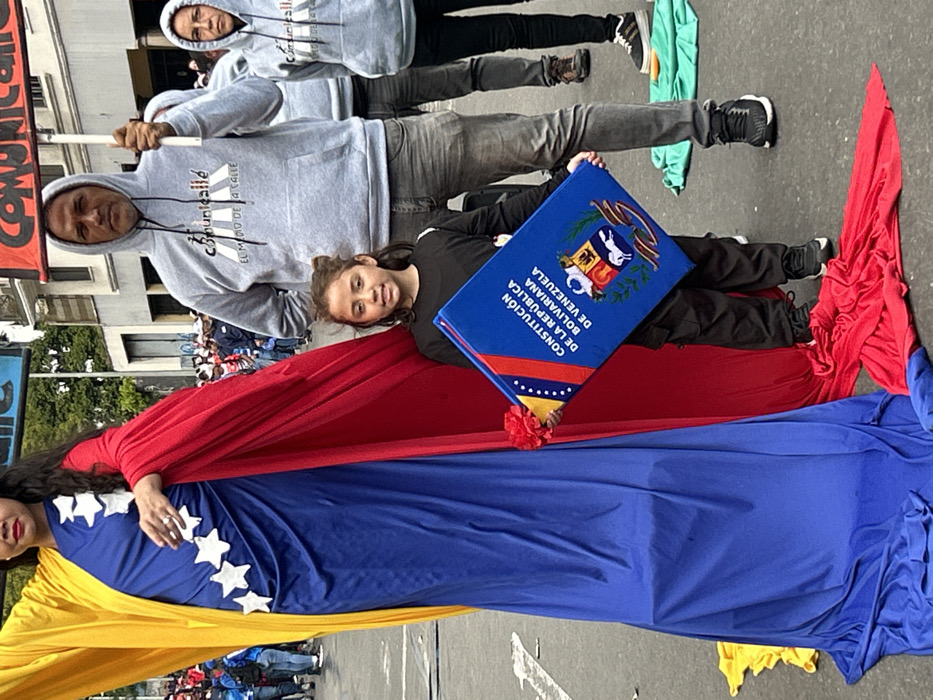 A group of people are seen at what appears to be a political demonstration. They are holding a large Venezuelan flag. A woman and a young girl are draped in the flag's colors. The girl is lying down and holding what looks like a constitution book, while the woman is also lying down, partially covered by the flag. Two men wearing hoodies are behind them, one carrying a sign with text.