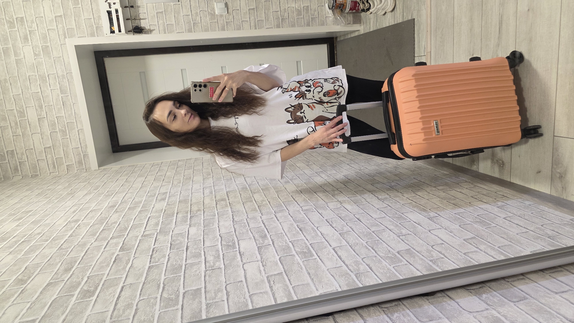 A woman with long, dark, wavy hair takes a selfie in a hallway with a light gray brick-patterned wallpaper. She is wearing a white t-shirt with cartoon dogs printed on it and black leggings. In her right hand, she holds a smartphone, and in her left hand, she grips the handle of a small, orange suitcase with wheels. The suitcase is positioned on the floor next to her. Behind her, a white door is visible, and to the right, a shelf with keys hangs on the wall. The floor is made of light-colored wood planks. The scene appears to be an indoor residential setting, likely a hallway or entryway in a home.