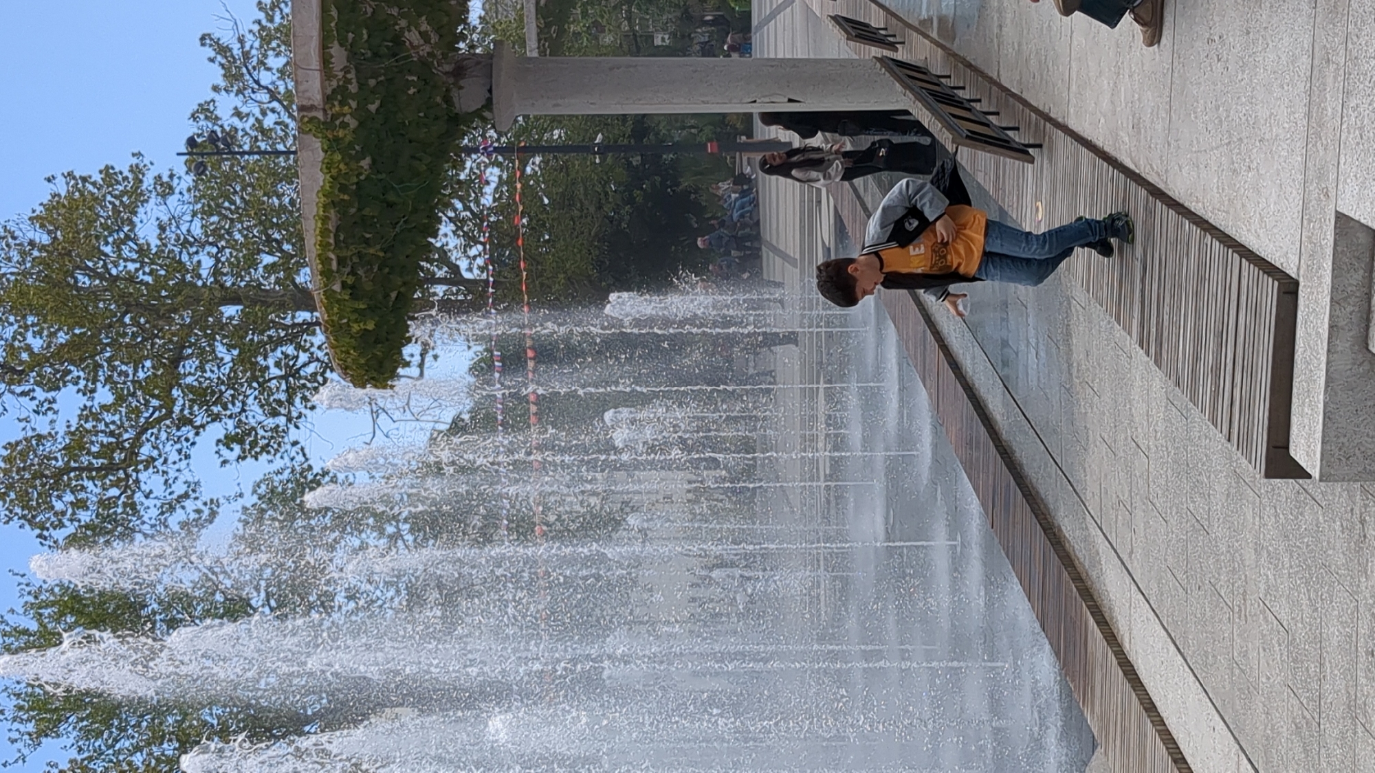 The image depicts a scene at a public fountain in the city of город Ялта, Ukraine. A boy is standing at the edge of the fountain, near a bench. Multiple streams of water are shooting upwards from the fountain, creating a dynamic water display. The background shows trees and greenery, suggesting a park-like setting. Other people can be seen in the distance behind the fountain. The overall scene suggests a public space for recreation and leisure.