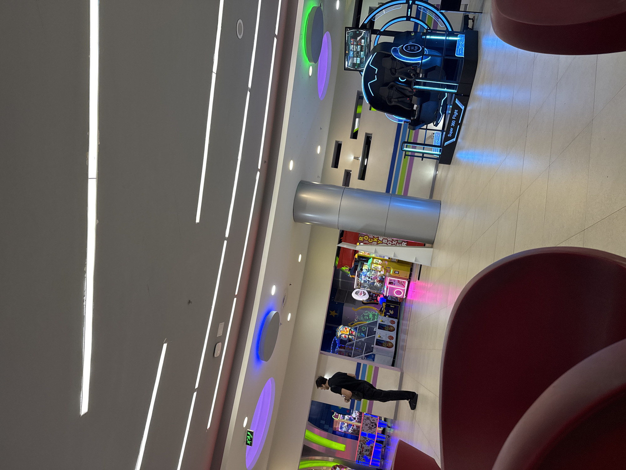 This media file depicts an indoor scene within a modern entertainment center or arcade, likely situated in a mall in Yerevan, Armenia. The space is brightly illuminated by numerous artificial light sources, including long, linear fixtures embedded in the ceiling and circular lights that cast green, purple, and blue hues. The floor is tiled with light-colored squares.
The central focus of the scene is an array of amusement rides and arcade games. A prominent feature is a "Super 360 Flight" virtual reality or motion simulator ride, characterized by its sleek black and blue design and two passenger seats. Beyond this, a collection of colorful arcade machines, including what appears to be claw games and a game with partial text "ROCK... BOXER," line the walls. Decorative elements like striped walls and a large silver cylindrical pillar contribute to the contemporary aesthetic.
A single individual, a man dressed in a black shirt and black trousers, is visible standing near the arcade machines. He appears to be observing the games or waiting, his presence indicating engagement with the leisure activities offered. In the immediate foreground, a portion of a large red chair is visible, suggesting a seating area for visitors. The overall atmosphere is one of modern recreation and entertainment. A green exit sign symbol is also noticeable on the ceiling.
