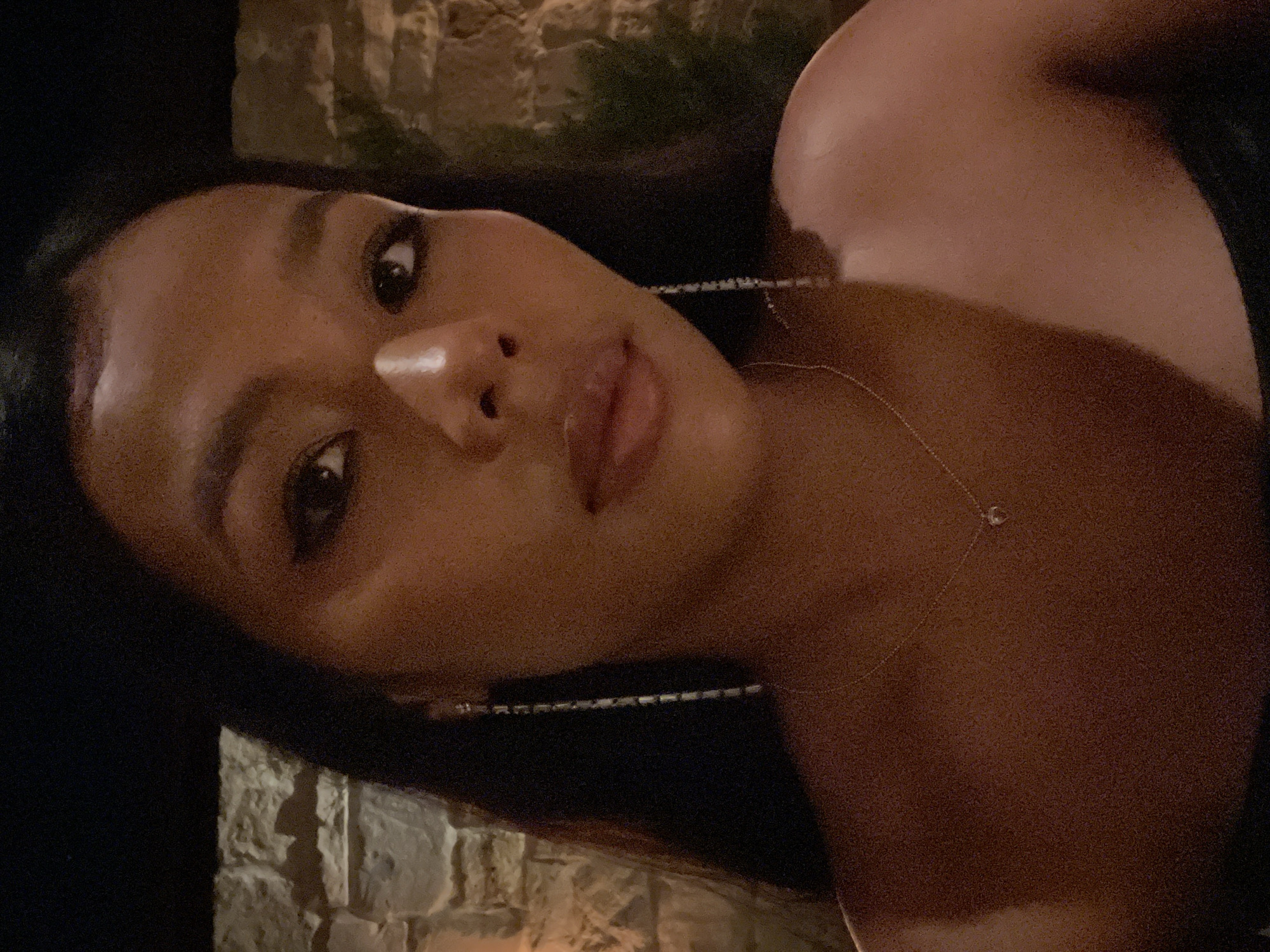 A close-up, waist-up, selfie-style photograph shows a woman with dark hair and a medium skin tone. She is wearing long, dangling earrings and a delicate necklace with a small, heart-shaped pendant. Her left shoulder and collarbone are visible. The background appears to be an outdoor setting with a stone wall and some foliage, suggesting a restaurant or outdoor event space. The lighting is dim, likely from artificial sources, creating a warm, intimate atmosphere. The image is set in North Kuta, Indonesia.